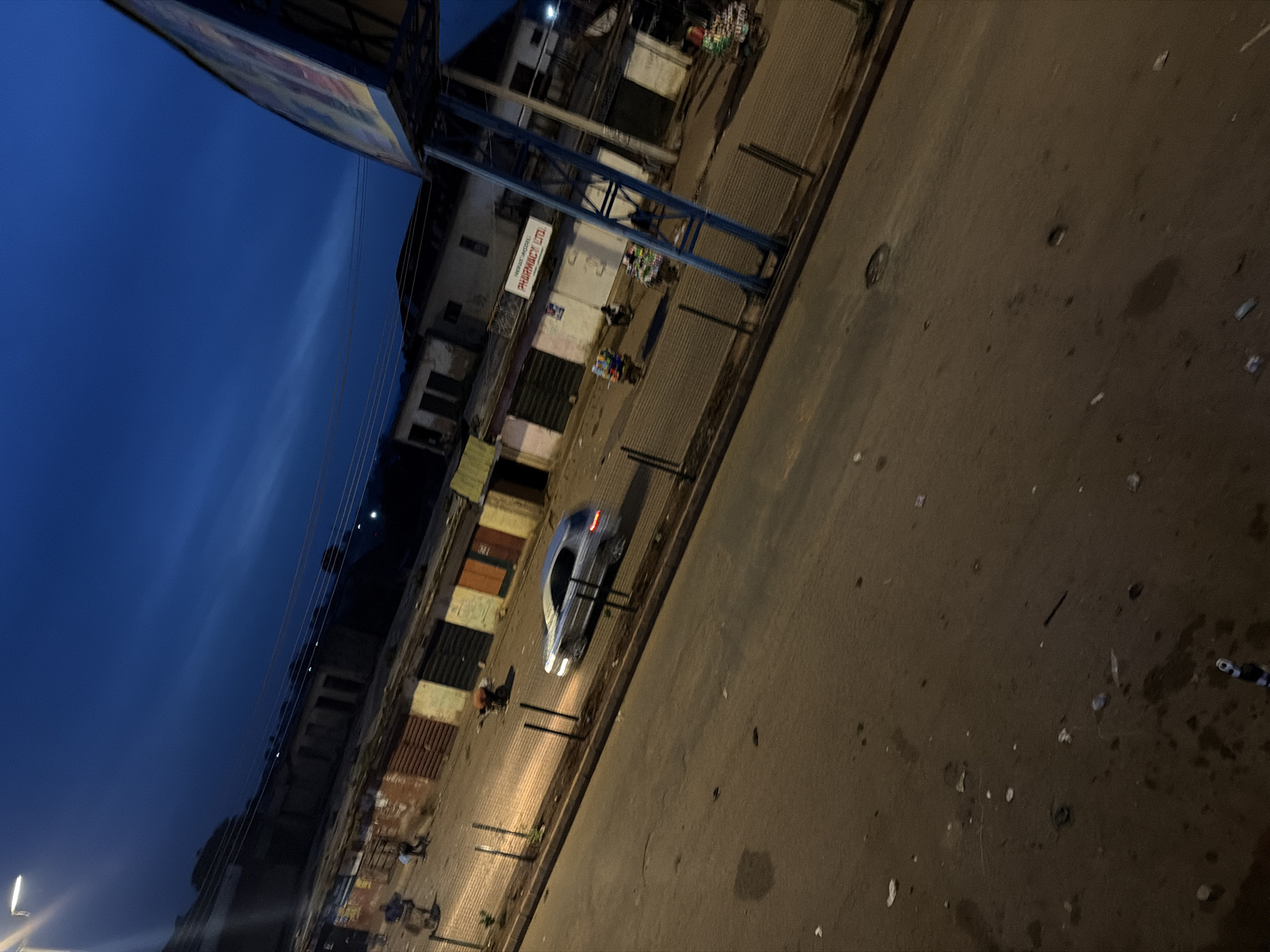 The image shows a street scene at dusk in Bukuru, Nigeria. A silver car is driving on the road, and there are several market stalls and buildings lining the street. People are visible in the distance, some of whom appear to be riding motorcycles or pushing carts. The sky is a deep blue, indicating twilight. The road surface is paved but appears somewhat worn and contains scattered debris. A large blue structure, possibly a billboard or a bus stop, is prominent on the right side of the frame. One building has a sign that reads "PHARMACY LTD." The scene appears to be a bustling commercial area.