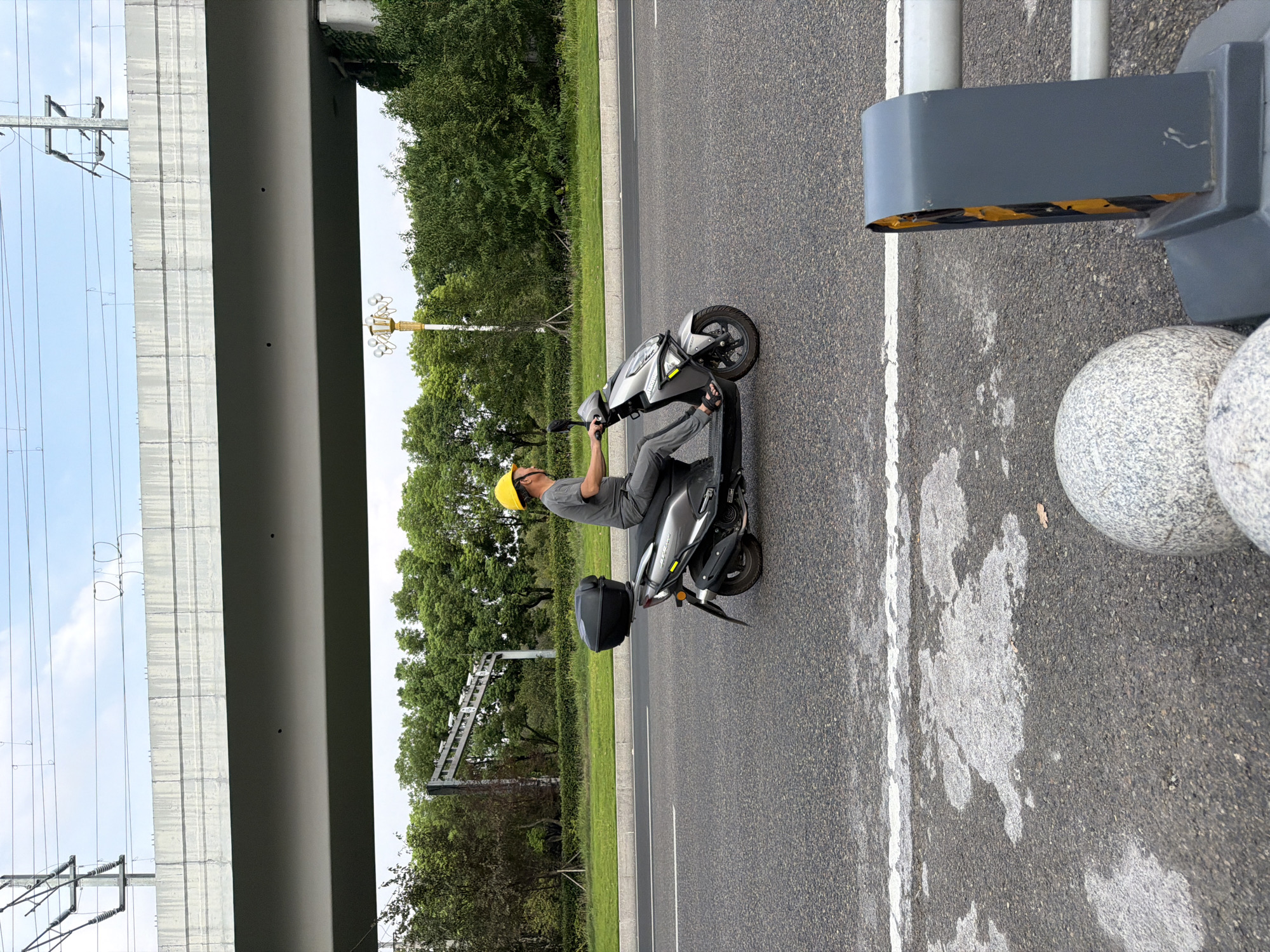 A man wearing a yellow hard hat rides a silver scooter with a black storage box on the back, traveling on an asphalt road in the city of Jiangjin, China. He is wearing a gray t-shirt and gray pants, and sandals. The road has white lane markings and appears wet in some patches. In the background, there is a large concrete overpass, lush green trees, a decorative lamppost, and power lines. The sky is overcast with some clouds. In the foreground, there are two large, round, gray, granite-like decorative spheres and a grey and yellow traffic barrier.