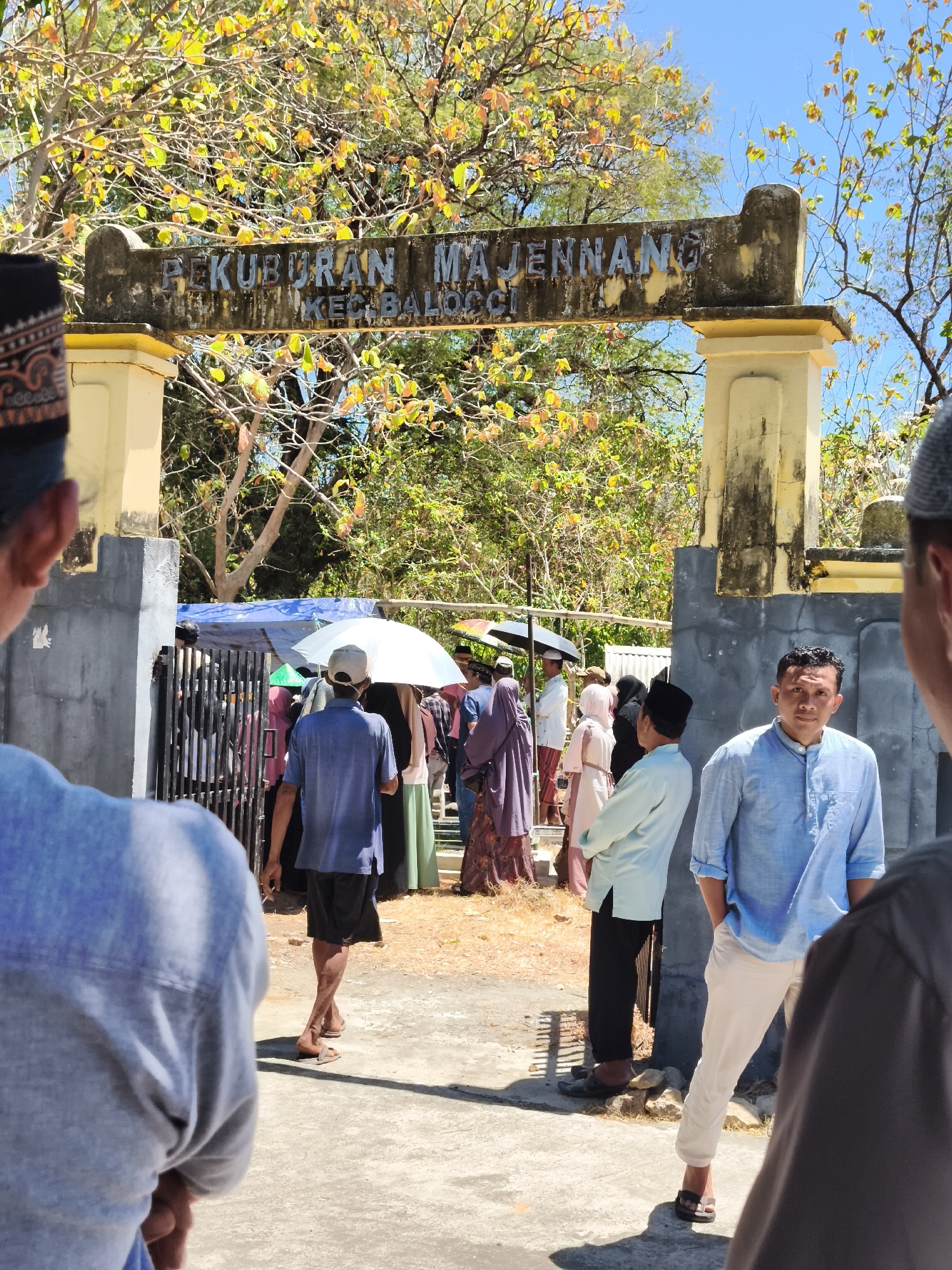 The image captures an outdoor scene in Tonasa, Indonesia, likely a cemetery entrance due to the inscription on the archway above. The arch reads "PEKUBURAN MAJENNA" and "KEC.BALOCCHI", identifying it as Majenna Cemetery in Balocci District. Several people are present, some gathered near a fence, while others stand in the foreground.
In the immediate foreground, the back of a person wearing a blue shirt and a patterned cap is visible, facing away from the camera. To the right, a man in a light blue long-sleeved shirt and cream-colored pants stands facing the camera, looking directly at it with a neutral expression. His hands are in his pockets.
Behind these figures, through an opening in a fence, a crowd of people can be seen. Some are holding umbrellas, one white and one possibly black, suggesting protection from the sun. People are dressed in various traditional Islamic attire, including hijabs and long garments in muted colors. One man in the middle ground wears a blue shirt and dark shorts and is walking away from the camera.
The environment appears to be dry and sunny, with a clear blue sky. Trees with yellowing leaves are visible behind the archway and around the entrance, indicating a possibly dry season or a specific type of foliage. The ground is a mix of concrete and dirt. The overall atmosphere suggests a gathering, possibly for a funeral or a visit to the cemetery.