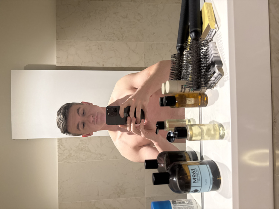 An adult male with short dark hair, appearing shirtless, is captured in a mirror selfie within a bathroom setting. He holds a dark-colored smartphone horizontally with both hands, directing his gaze downwards towards the device. The background features light-colored, marble-patterned tiled walls and a reflective white counter surface. On the counter, various toiletries are arranged. From left, these include a blue aerosol can, two dark-colored pump bottles, a clear amber bottle with a blue label displaying "M061 PERFUME LAB 3 x 10 ml Venezia," a smaller clear bottle with a black pump, another amber bottle, a white cylindrical container, two black-handled hairbrushes, and a bar of soap. A large rectangular mirror reflects part of the ceiling above the subject. The presence of the label "Venezia" on one of the bottles indicates the location is Venice, Italy.