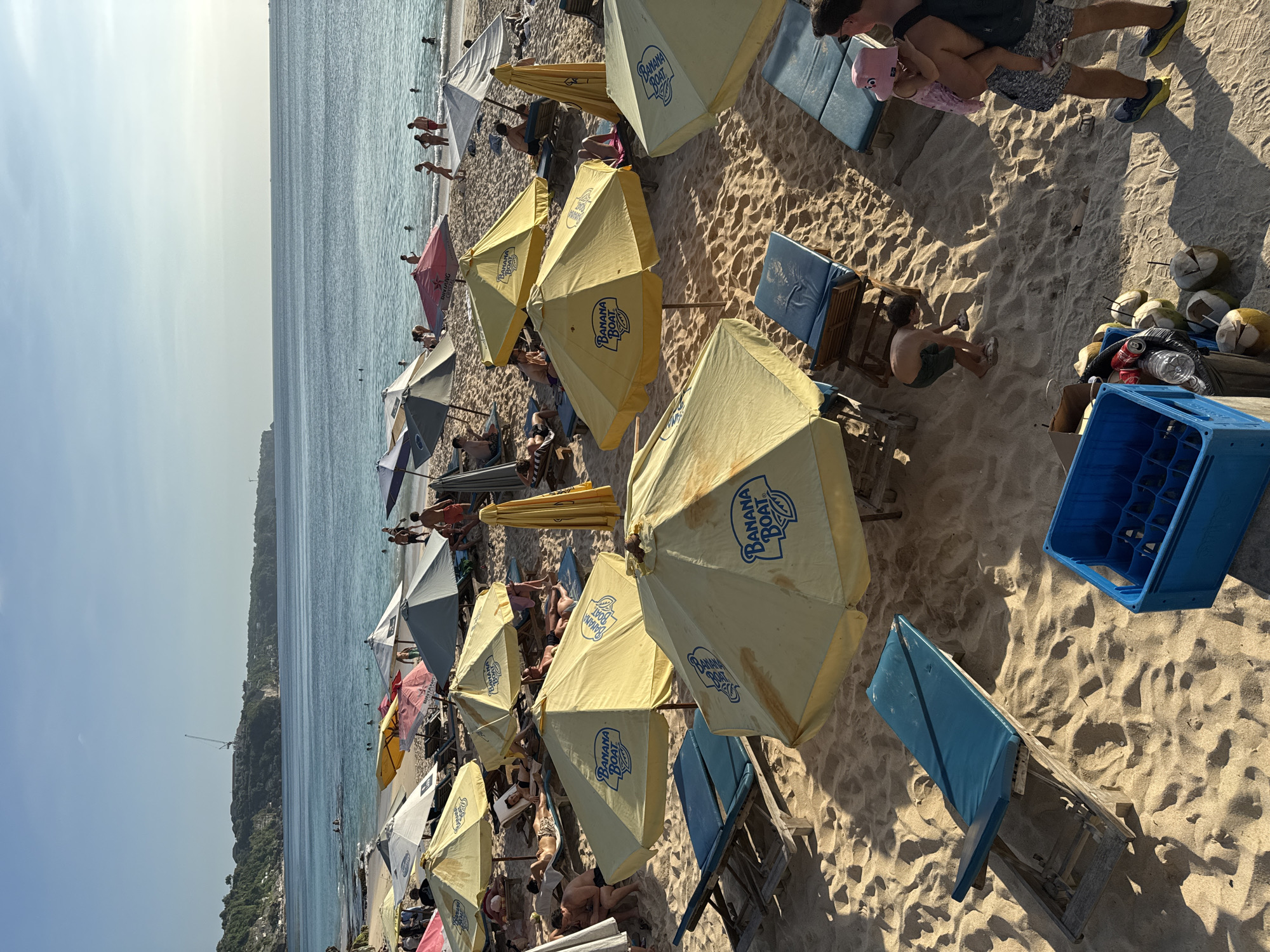The image shows a densely populated beach in Kuta Selatan, Indonesia, filled with numerous beach umbrellas, many adorned with the "Banana Boat" logo. Beachgoers relax under the umbrellas on lounge chairs. In the foreground, a blue plastic crate and a collection of coconuts and discarded beverage containers are visible. Further along the beach, people are seen swimming and interacting with the water. A dense green cliff is visible in the distance behind the beach. The sky is clear and light blue.