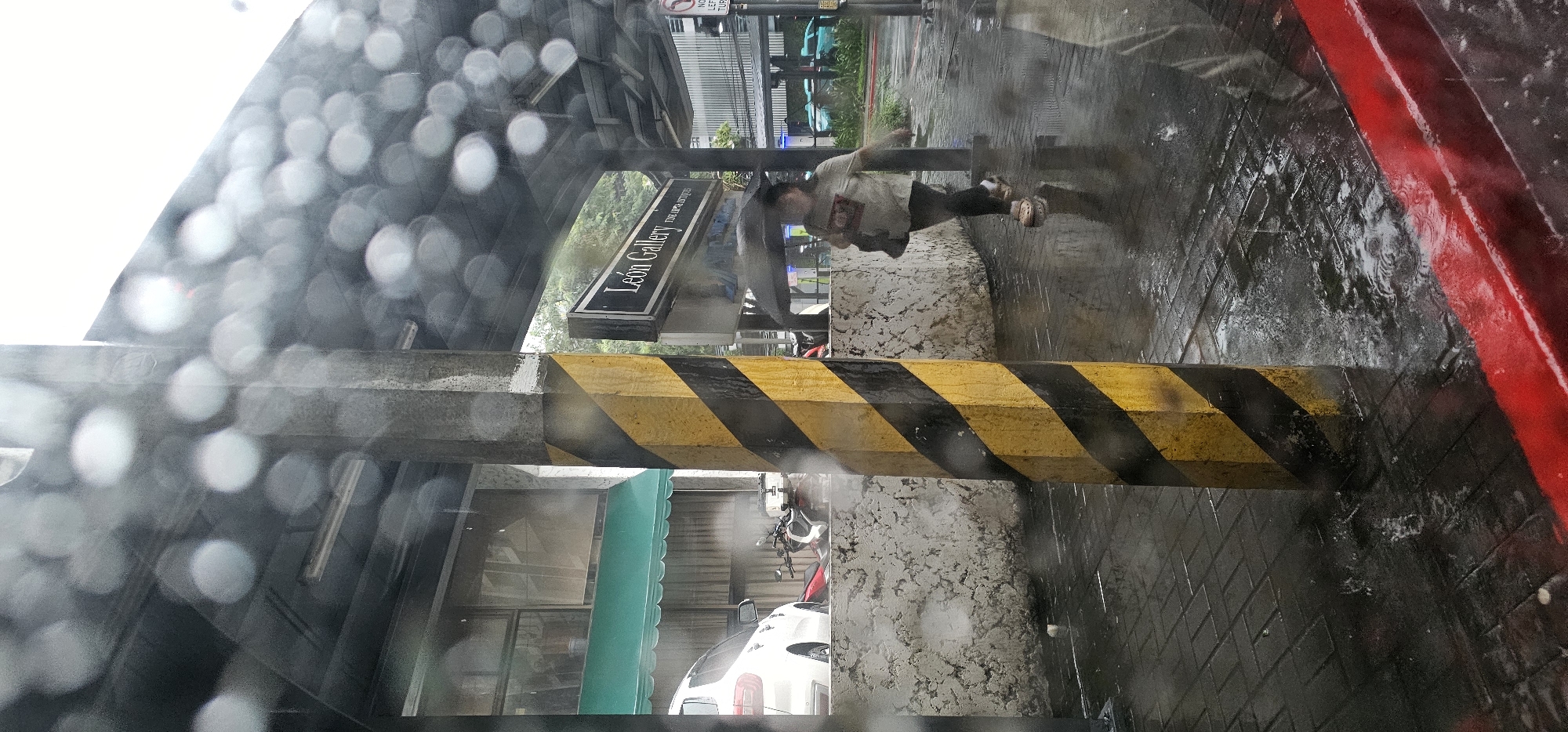 The image depicts a street scene in Makati City, Philippines, during a rain shower. A pedestrian, a person of indeterminate gender wearing a light-colored shirt, dark pants, and sandals, is walking under a dark umbrella. The person is moving from right to left across the frame. To the left of the pedestrian, a concrete pillar with diagonal yellow and black stripes stands prominently. Behind the pillar, a white SUV is parked. Further back, a building with a green awning and windows is visible. A sign that reads "León Gallery" is mounted on the right side of the frame, indicating the presence of an art gallery. The ground is wet and reflective, with visible puddles and ripples from the rain, suggesting it is a paved area, likely a sidewalk or street. The overall environment is urban, characterized by the presence of buildings, vehicles, and street infrastructure. The visual quality is somewhat obscured by water droplets on the camera lens or viewing surface, creating bokeh effects.