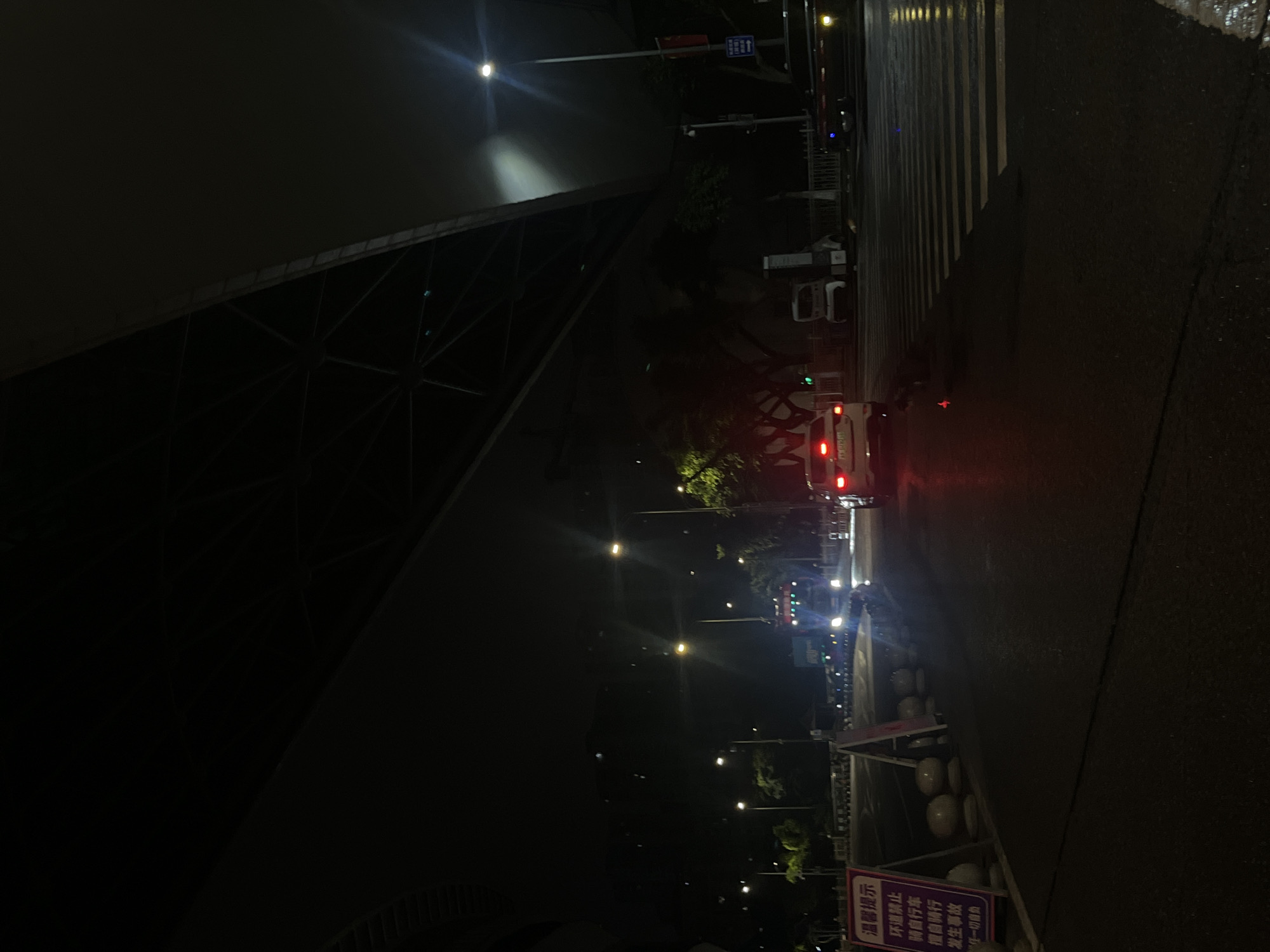 The image depicts a nighttime urban scene under a large, dark overhead structure, likely an elevated roadway or bridge, within Jiulongpo District, China. The asphalt road is visibly wet and reflective, suggesting recent precipitation.
In the foreground and mid-ground, a white-striped pedestrian crossing is prominent on the road surface. On the left side of the road, a purple and white sign displays Chinese characters, "温馨提示" (Warm Reminder), followed by instructions pertaining to prohibited bicycle use and individual responsibility for accidents. This sign is situated near a line of white spherical bollards and other barriers.
Multiple vehicles are present. A blue truck with its headlights illuminated is observed approaching from the center-left mid-ground. On the right side of the road, a white sedan displays illuminated red taillights, facing away from the viewer. Further in the distance, additional vehicle lights indicate ongoing traffic. A small white utility vehicle is parked on the far right side of the road, adjacent to what appears to be urban infrastructure.
The environment is illuminated by numerous streetlights emitting both white and yellowish light, casting reflections on the wet road. Green foliage from trees is visible along the roadside, partially obscured by the darkness and overhead structures. No identifiable human figures are present in the frame.