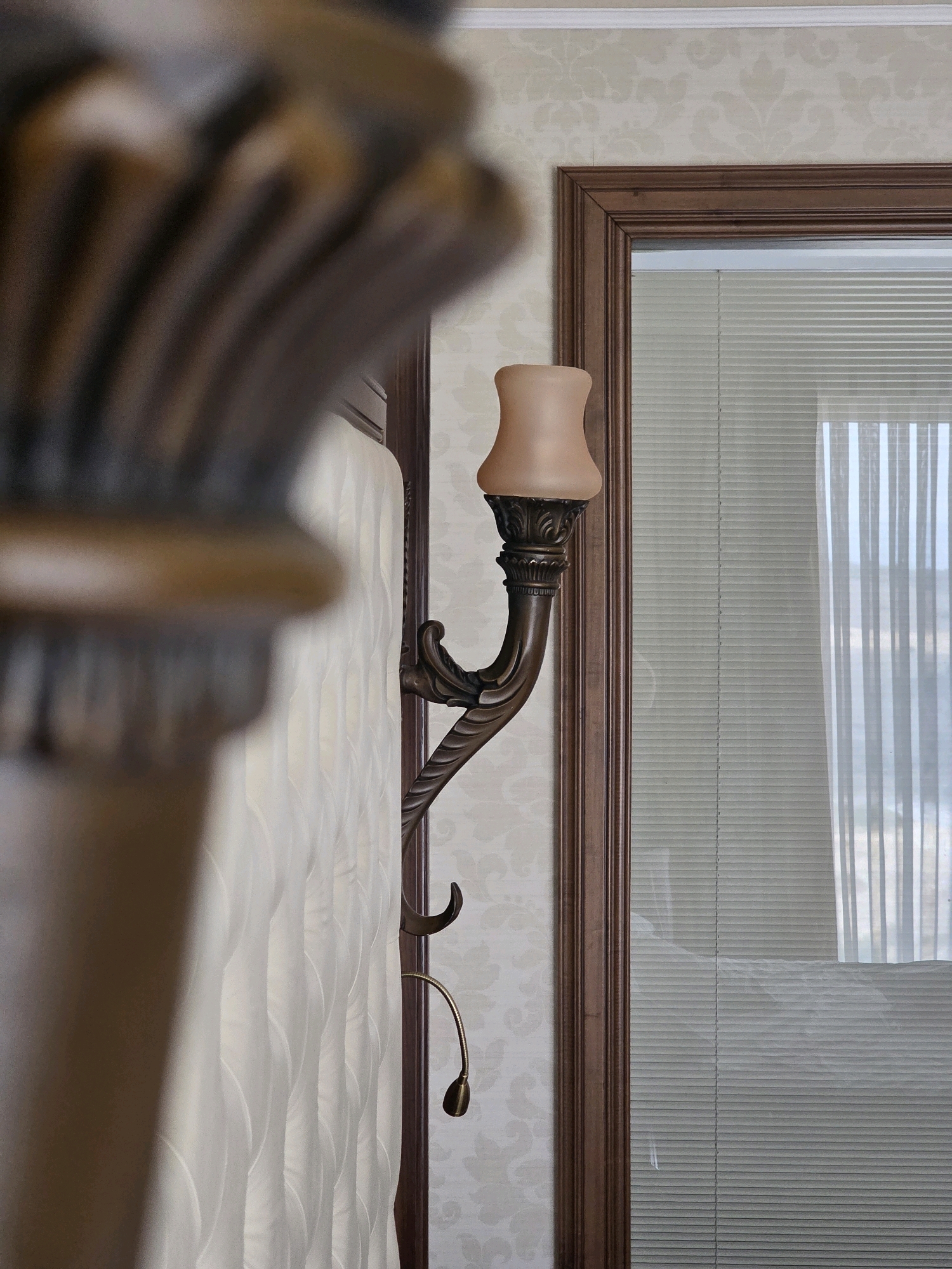 The image depicts an indoor scene with a focus on decorative elements. In the foreground, a blurred wooden object, possibly part of a headboard or furniture, occupies the left side of the frame. To the right, a wall-mounted sconce with an ornate bronze finish and a peachy-colored glass shade is visible. This sconce is attached to a wall covered in a subtle damask-patterned wallpaper. Adjacent to the sconce is a dark wood frame, likely surrounding a mirror or a window. Inside this frame, horizontal blinds are visible, with vertical light reflections suggesting a source of light from outside. A small, adjustable reading lamp with a brass finish extends from beneath the sconce. The overall aesthetic suggests a bedroom or a similar personal space with a classical or traditional design.