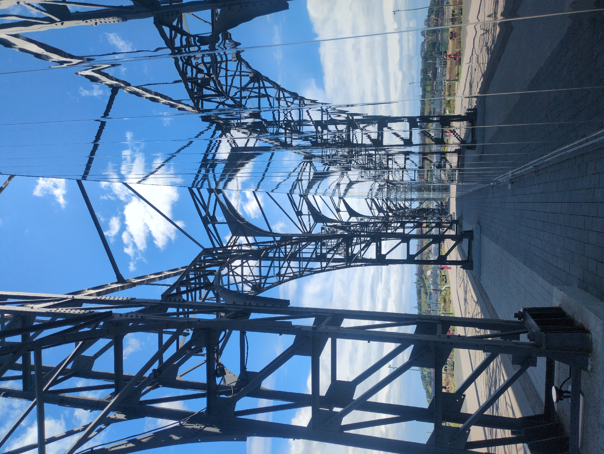 The image depicts the Nizhny Novgorod Kremlin's "Nizhny Novgorod Sky" observation deck, a contemporary art installation. The structure is characterized by a complex metal framework, resembling industrial scaffolding, that supports a polished, reflective surface on one side. This reflective surface distorts and mirrors the sky, the clouds, and the metal framework itself, creating a surreal effect.
The foreground features a paved walkway made of gray interlocking bricks. On the left side of the walkway, a large section of the dark metal structure rises. Its beams and supports are intricately interconnected, forming an abstract, arching shape that recedes into the distance.
To the right of the walkway, the highly polished, vertical reflective panels form a wall that extends into the background. These panels capture the sky and the metalwork, breaking them into fragmented, wavy lines. The sky above is a bright blue, punctuated by scattered white clouds.
In the middle ground, along the walkway, several people are visible. A person in a pink outfit walks away from the camera towards the left. Further down the walkway, two individuals are seen on the right side, and one person in an orange outfit is walking towards the left. Beyond the walkway, a grassy area with sparse vegetation is visible, leading to a distant cityscape with buildings and green hills under a bright sky. The overall impression is one of a modern urban space designed to interact with its natural and architectural surroundings through visual distortion and reflection.