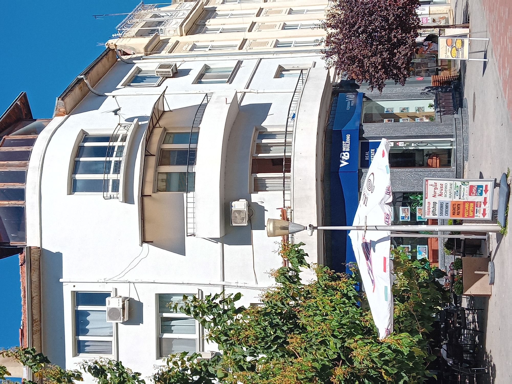 The image depicts a street scene in Varna, Bulgaria, focusing on the facade of a white building with a curved corner. The building has multiple stories with numerous windows, some featuring balconies with black railings. An air conditioning unit is visible on one of the exterior walls. Below the white building, a blue storefront with "WEST BUILDING" signage is present, indicating a business establishment.
In the foreground, a white patio umbrella shades a seating area with tables and chairs, suggesting an outdoor cafe or restaurant. A streetlamp stands next to the umbrella. To the right of the umbrella, a sign displays various items for sale, including "giftshop," "souvenirs," and "postcards." Further to the right, a person is walking on the sidewalk, and another sign displays images of food.
To the right of the main white building, another building with a lighter facade and arched windows is visible, hinting at older architecture in the vicinity. The sky is clear and blue, and sunlight casts shadows on the buildings and ground. A tree with dark foliage stands to the right of the storefront. A person with a backpack is walking on the sidewalk near this tree.