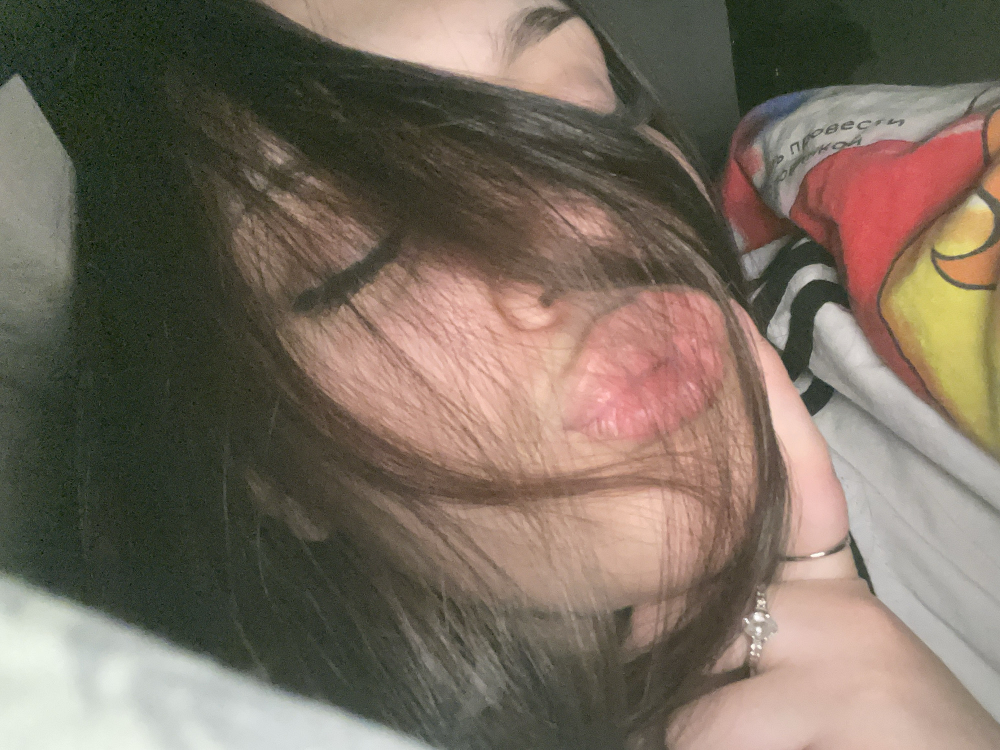 The image is a close-up shot of a person lying down, possibly on a bed. The person has dark hair that partially covers their face, with a focus on their lips. The lips are a prominent pink color. Their eyes are closed. The person's right hand is visible with rings on the fingers. The background includes a pillow with a printed design in Russian, and a striped sheet. The environment appears to be indoors, possibly a bedroom. There is no definitive information to determine if the location is Murino, Russia.
