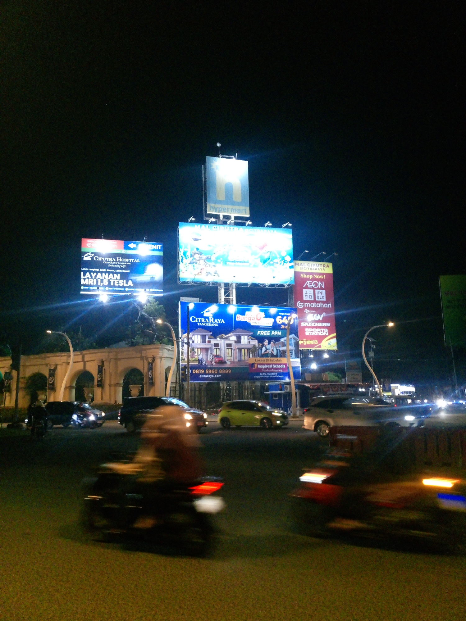 The image shows a night scene with multiple illuminated billboards at an intersection in Bitung Jaya, Indonesia. The billboards advertise various businesses including Ciputra Hospital, Mal Ciputra, and apartments with "Lokasi di Sebelah" (Located next door). One billboard for Ciputra Hospital highlights "Layanan MRI 1,6 Tesla" (MRI 1.6 Tesla Service). Another prominent billboard features logos for AEON, UNIQLO, Matahari, and SKECHERS. The street is busy with traffic, including cars and motorcycles, with motion blur indicating movement. Streetlights and building lights contribute to the overall illumination of the scene. In the foreground, two motorcycles are in motion, with the rider and vehicle of the closest one blurred due to the exposure. The background features buildings with arched structures and trees. The sky is dark, indicating nighttime.