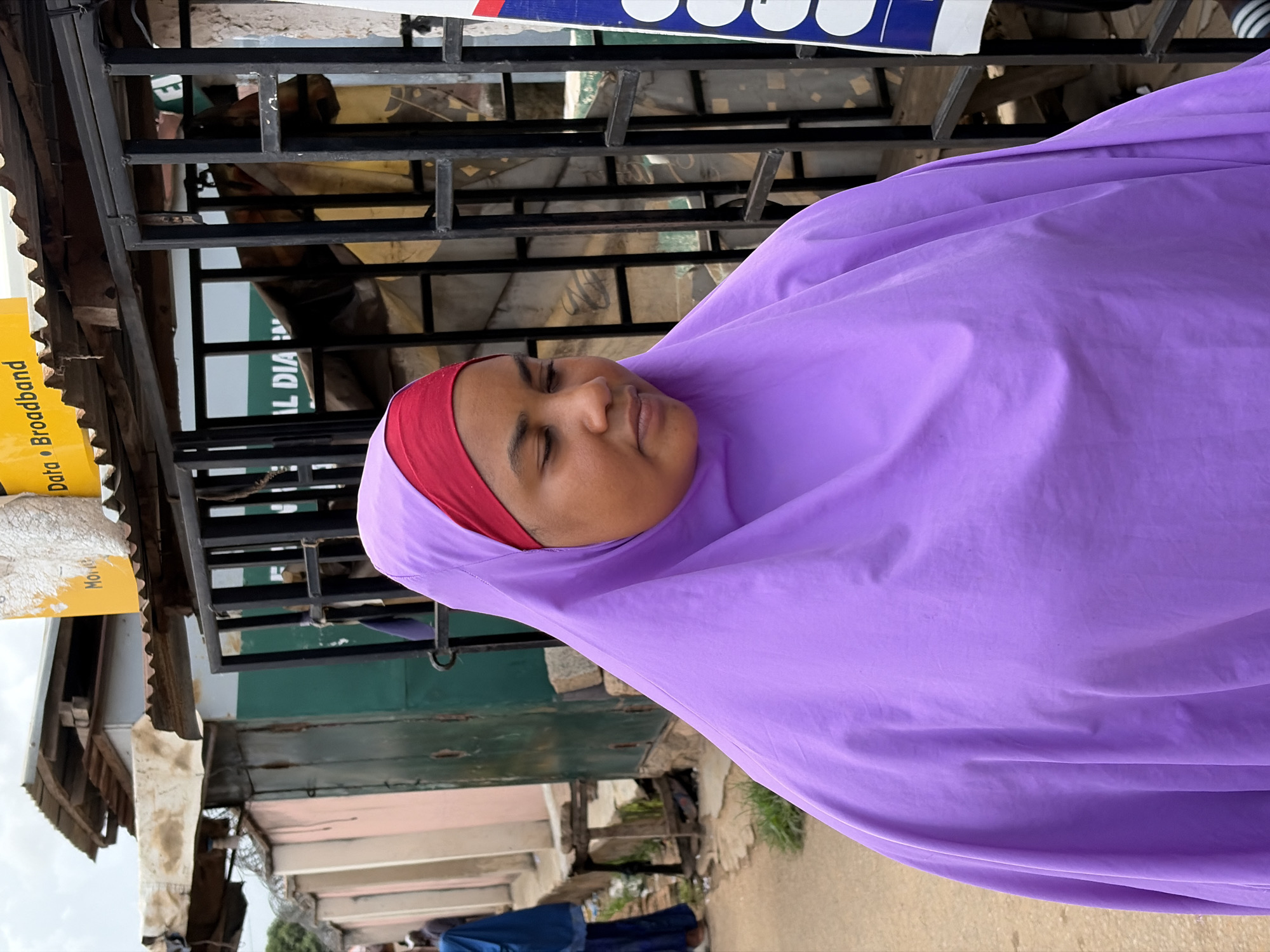 An outdoor, eye-level shot, rotated 90 degrees clockwise, captures a woman with her eyes closed, seemingly resting or in a contemplative state. She is wearing a vibrant purple hijab and a visible red underscarf. Her face is in focus, showing a calm expression.
The background, partially obscured by the woman's purple garment and a black metal grating structure, suggests a bustling street or market environment in Jos, Nigeria. Through the grating, glimpses of other people and rudimentary structures can be seen. On the far left, corrugated metal roofs and yellow signs are visible. One yellow sign clearly displays "Data" and "Broadband," with a partially visible "Mone" which could indicate "Money" or "Mobile Money." Another sign, further back and green, has partially visible text "IAL DIA". At the top, a blue and white sign with circular elements is present, though its full text is not decipherable.
The lighting indicates it is daytime, casting even light on the scene. The overall impression is a candid moment captured in an urban or semi-urban setting, characterized by local shops or stalls.