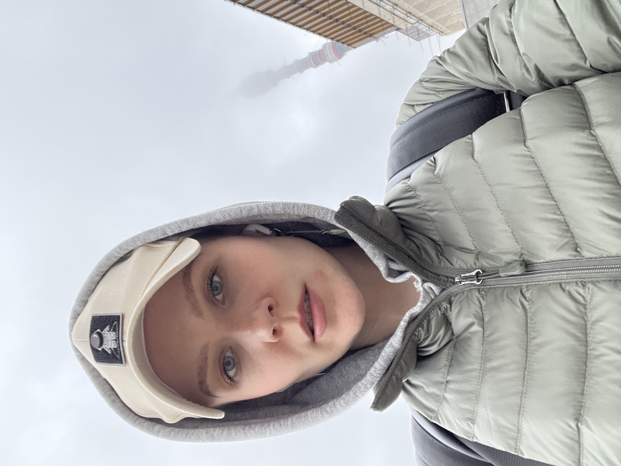 A young woman with fair skin and blue eyes is centered in the frame, looking at the camera. She is wearing a light-colored baseball cap with a logo, a grey hooded sweatshirt, and a light green puffer jacket. A black strap is visible across her right shoulder.
Above her, out of focus, is a tall structure which is partially obscured by fog or clouds. The structure appears to be a tower with red and white bands around it. Some construction scaffolding is also visible. This is the location in the city of Khimki, Russia.