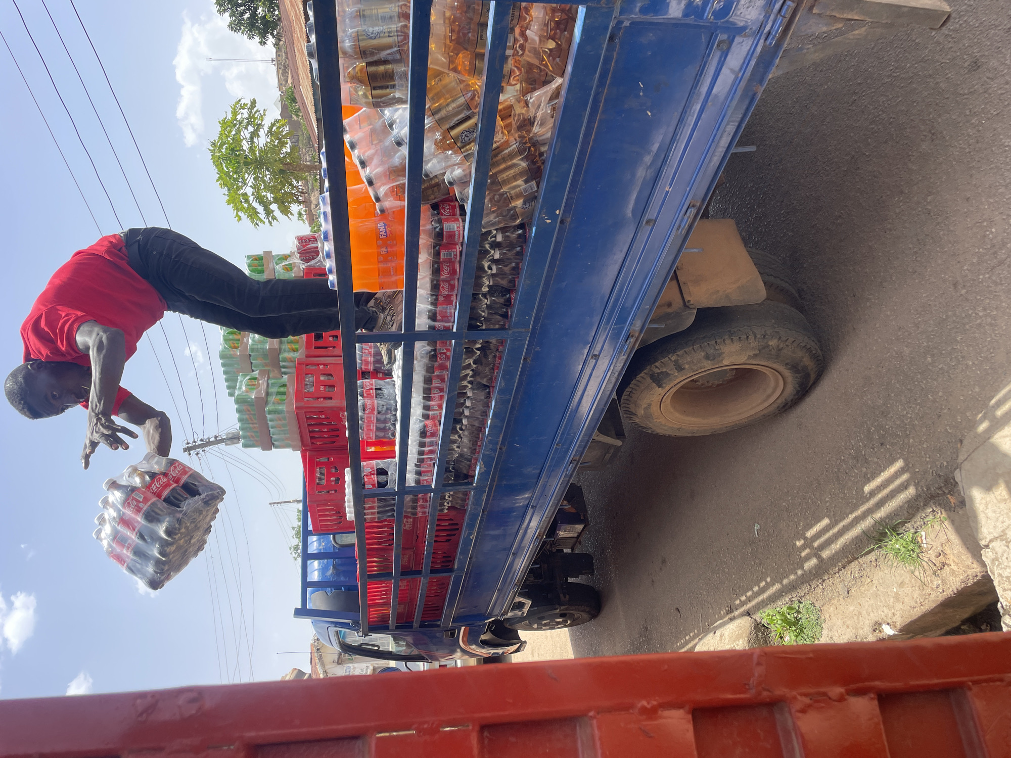 A man is unloading bottled beverages from the back of a blue truck in Jos, Nigeria. The man, wearing a red shirt and black pants, is bending over to hand a plastic-wrapped package of Coca-Cola bottles. The truck bed is loaded with cases of Coca-Cola, Fanta, and other drinks. The scene is outdoors, and there is a tree visible in the background.