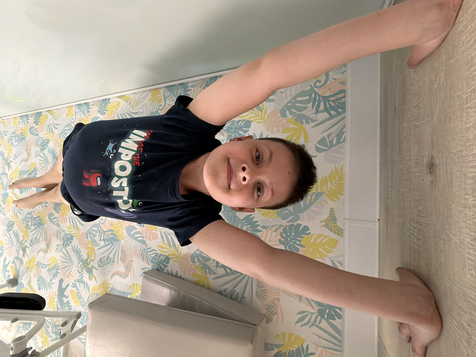 A young person with light skin, short dark hair, and wearing a navy blue t-shirt with "Among Us" graphics is lying on a bed with a patterned duvet cover. The pattern consists of stylized yellow and blue leaves. They are positioned with their head toward the top of the image and their feet toward the bottom left. The person has their arms outstretched, with their hands pressed against the wall and the edge of a piece of furniture. The furniture appears to be a white chair or ottoman. A beige wall is visible on the right side of the image. The location of the image is unknown and there is no evidence to suggest the location is the city of Votkinsk, Russia.