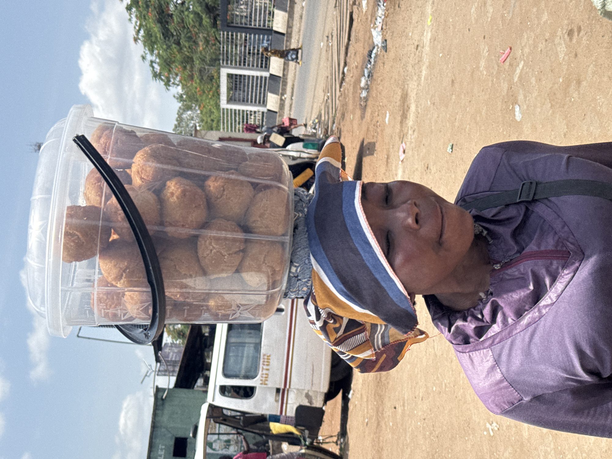 The image, taken in Onitsha, Nigeria, depicts a street vendor carrying a large clear plastic container filled with baked goods on their head. The main subject, likely a woman, is in the foreground, wearing a purple long-sleeved garment and a vibrant headscarf patterned in blue, white, yellow, and brown. A black strap, possibly from a bag, crosses her chest. Her face, visible in profile, shows a calm, neutral expression as she looks slightly upwards.
The transparent container, which has a black handle, is filled with numerous round, golden-brown baked items, resembling puff-puff or a similar fried dough snack, piled high. This method of carrying wares on the head is characteristic of street hawkers in the region.
The setting is an outdoor, bustling street scene during bright daylight, indicated by the clear blue sky with scattered white clouds. The ground is a sandy, reddish-brown dirt, showing some scattered litter. In the background, a white bus or commercial vehicle is partially visible, with the word "MOTOR" discernible on its side, along with other indistinct structures, fences, and greenery. Another person can be seen walking further down a paved road in the distance, underscoring the active, informal street commerce environment.