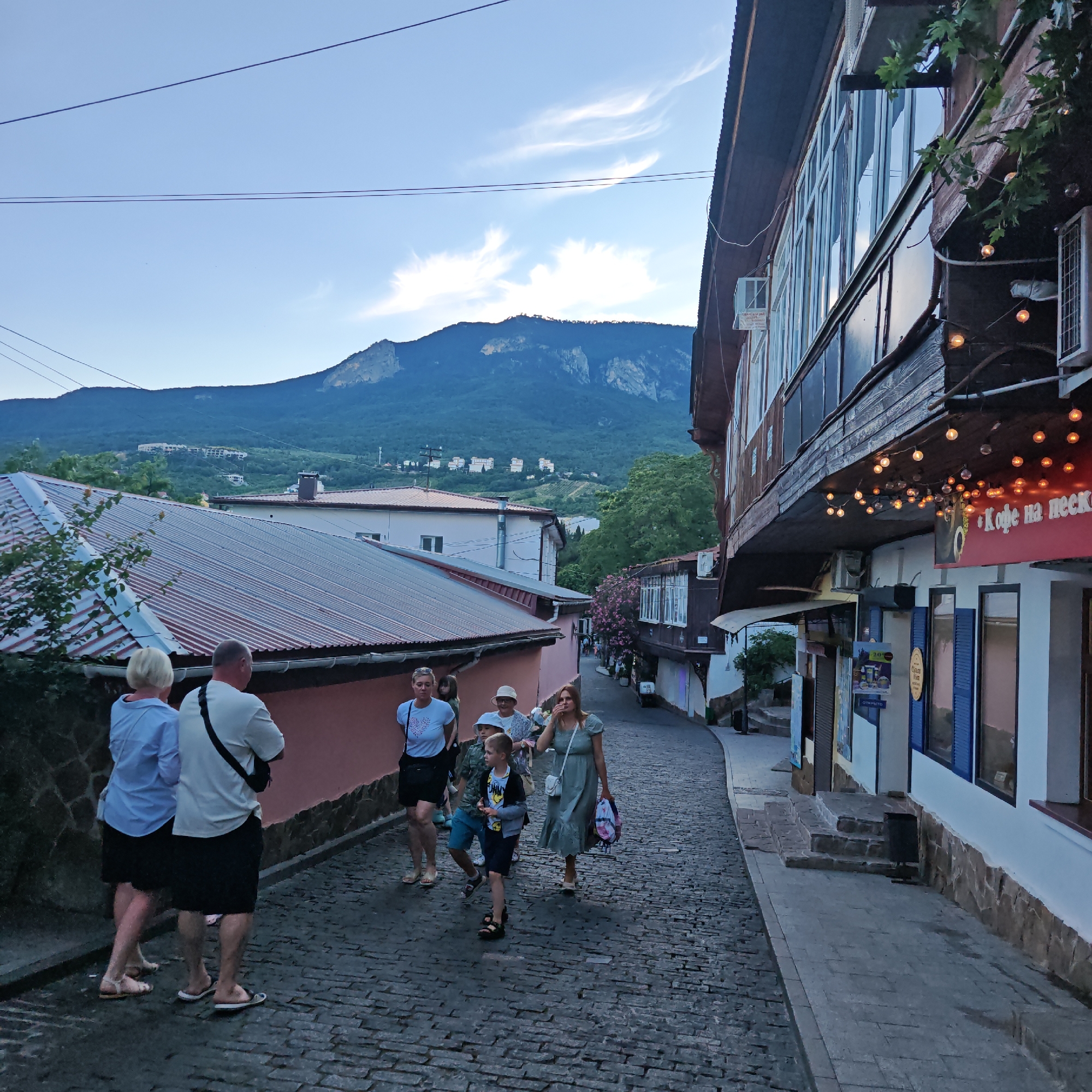 In Гурзуфский поселковый совет, Ukraine, a cobblestone street slopes gently uphill, lined with buildings and a mountainous backdrop. The evening sky is a dusky blue with wispy white clouds, and the sun has recently set, casting a soft, diffused light.
On the left side of the street, a cluster of people is walking uphill. A woman in a blue shirt and black skirt is in the foreground, her back to the camera. Next to her, a man in a white t-shirt and black shorts, with a black strap across his chest, is also walking away from the viewer. Behind them, a younger woman in a white t-shirt and black shorts walks with a young girl. Further up the street, a boy wearing a light-colored hat and a gray jacket over a t-shirt walks with another child.
Across the street, a building with a dark wooden facade features several windows and a balcony adorned with string lights. A sign with Cyrillic text, likely indicating a business, is visible. The building has a warm, inviting glow from the lights. Further down the street, more buildings are visible, including one with a reddish-brown corrugated metal roof.
The mountainous landscape dominates the background, with lush green trees covering the lower slopes and rocky peaks rising towards the sky. The overall scene conveys a sense of a relaxed evening stroll in a picturesque town.