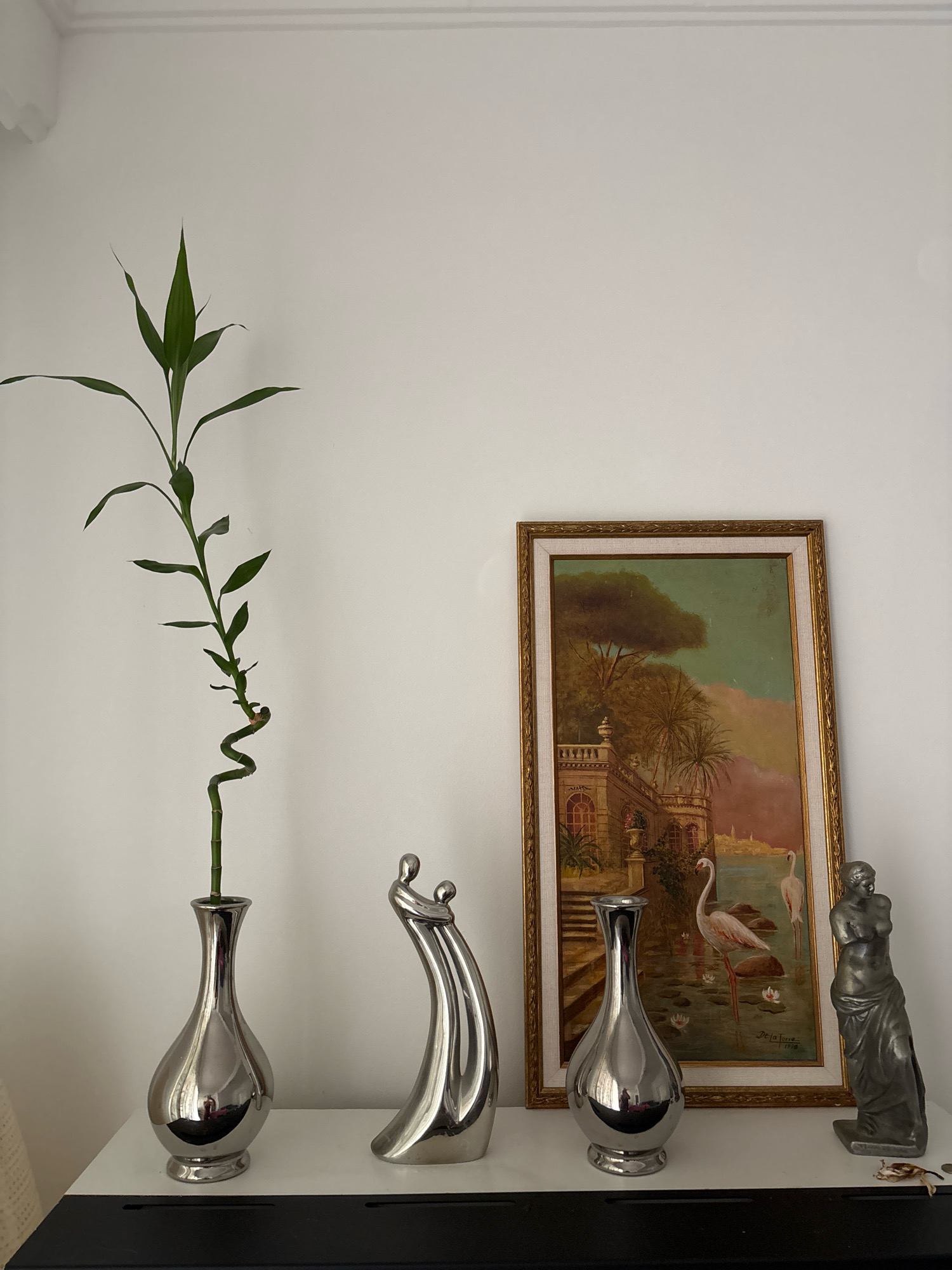 A still life arrangement is presented on a white shelf against a plain white wall. To the left, a tall, spiral-shaped Lucky Bamboo plant stands in a sleek, silver vase with a rounded base and a narrow neck. Next to the plant, a modern, abstract sculpture depicts two stylized human figures embracing, also in a metallic finish. To the right of the plant is another identical silver vase.
Dominating the center of the arrangement is a framed oil painting, leaning against the wall. The painting depicts a classical European scene with an ornate building, palm trees, and a body of water. Two white flamingos are visible in the water, along with lily pads and reflections. The painting is signed "De La Torre" and dated "1978".
To the far right, a dark gray statue of a classical female figure, possibly Venus, stands. The statue is draped in fabric and appears to be made of metal. A small, dried organic object, possibly a leaf or a dead insect, rests on the shelf in front of the statue. The overall scene suggests an indoor decorative display, likely in a home or gallery setting. The image does not provide any specific information to pinpoint a location in Malaga, Spain.