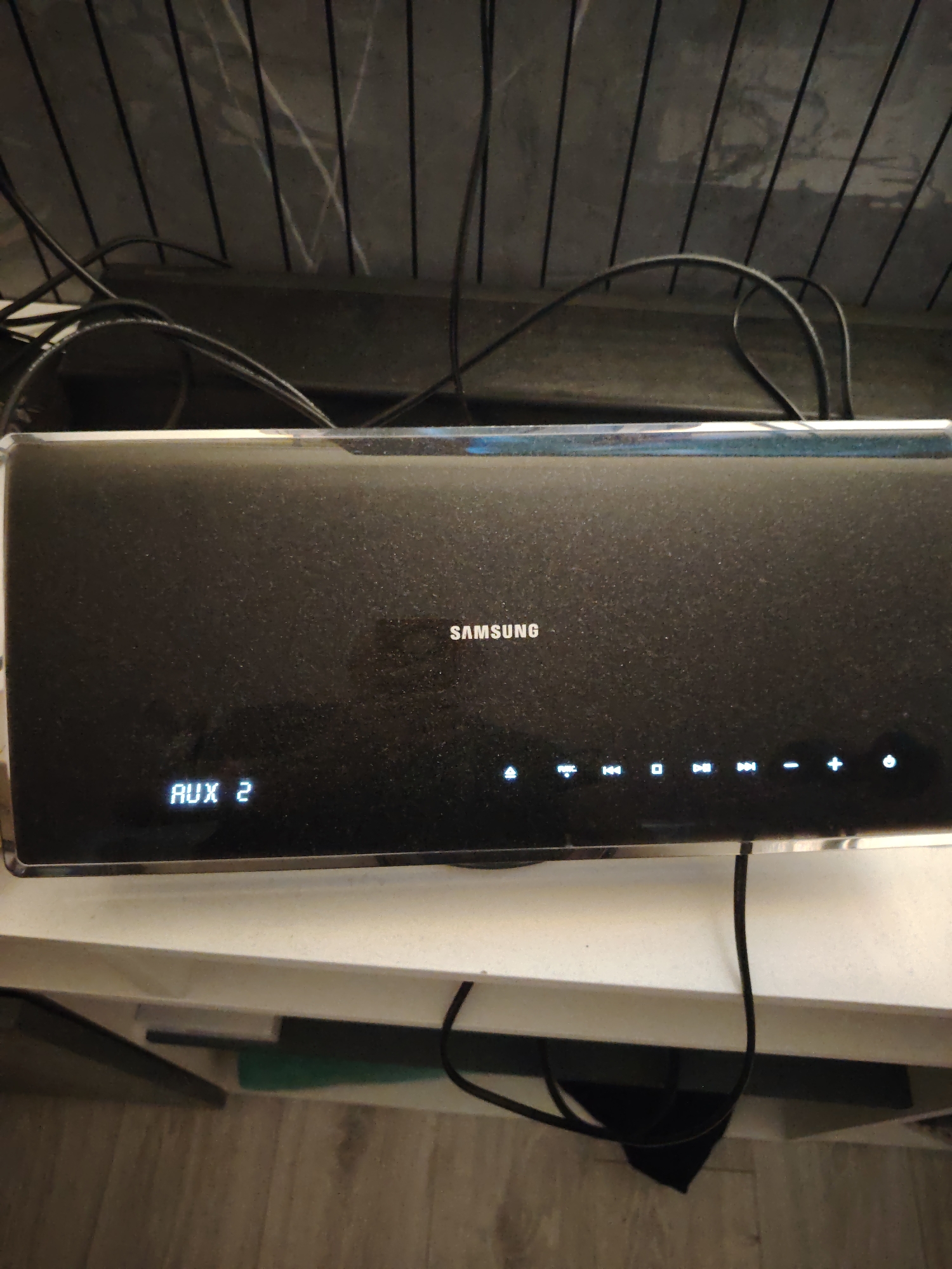 The image displays a black Samsung soundbar positioned on a white shelf, with various cables draped behind and beneath it. The soundbar's surface is visibly covered in dust particles. Its central display shows "SAMSUNG" in white lettering. To the left, "AUX 2" is illuminated in white, indicating the selected input source. To the right, a series of touch-sensitive or illuminated control icons are visible in white: a source/menu icon, "FUNC", rewind, stop, play/pause, fast forward, volume down (-), volume up (+), and a power button. Above the soundbar, a dark, textured surface with vertical linear patterns is visible, crisscrossed by multiple black cables. Below the white shelf, a light-colored wooden or laminate floor with a visible grain pattern extends from the bottom of the frame. The scene is located in Huntingdonshire, United Kingdom.
