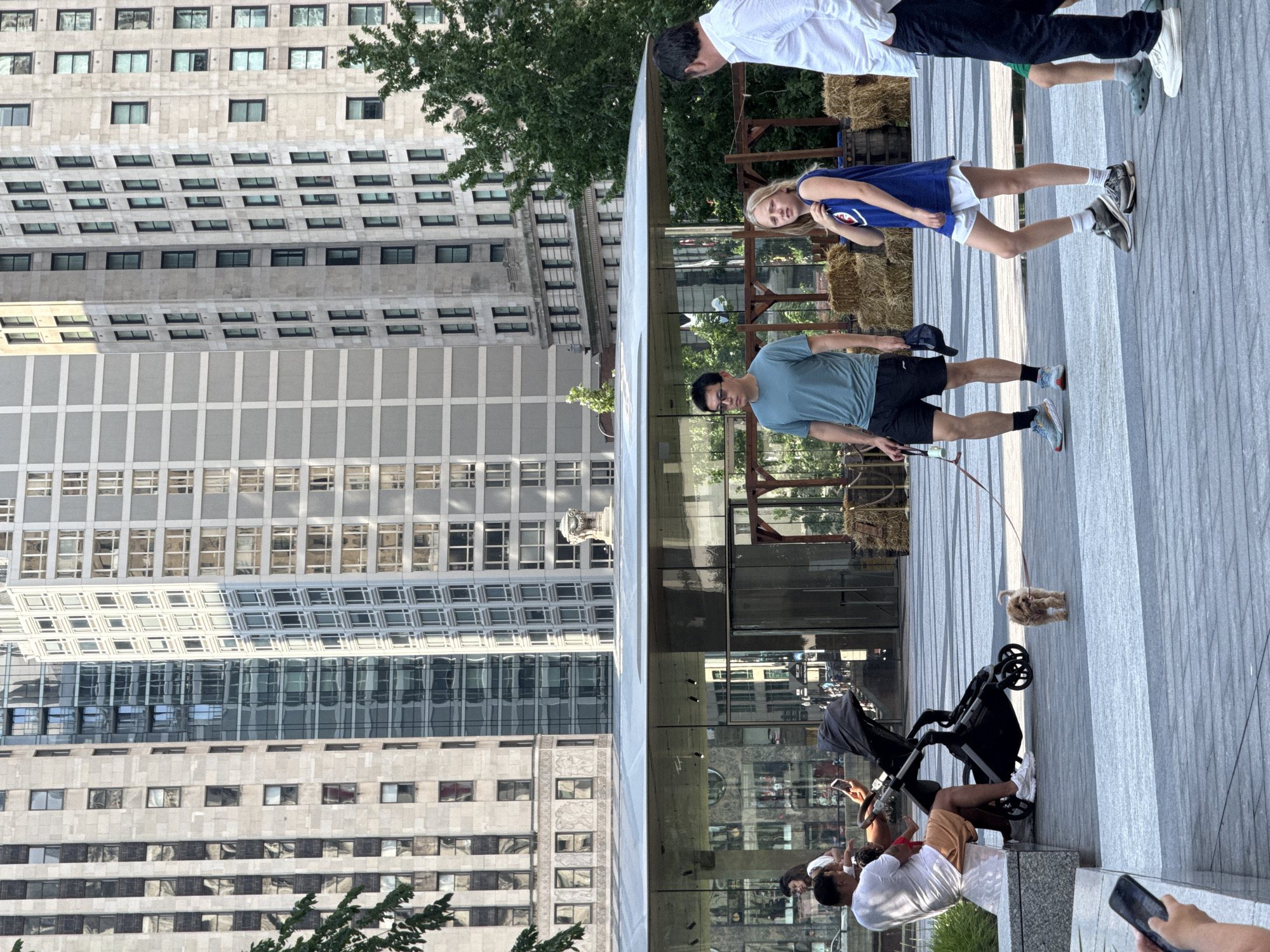 The image captures a scene in Millennium Park, Chicago, United States of America. In the foreground, a man in a blue t-shirt and black shorts walks a small, light-brown dog on a leash. Beside him, a young girl in a blue tank top and white shorts walks in the same direction, looking towards the viewer. To the far right, a man in a white shirt and black pants walks away from the camera. In the midground, a man in a white t-shirt and orange shorts sits on a bench next to a stroller, holding a smartphone. The background features a modern building with a glass facade, behind which stand several tall, imposing skyscrapers, characteristic of the Chicago skyline. The ground is paved with large, gray tiles.