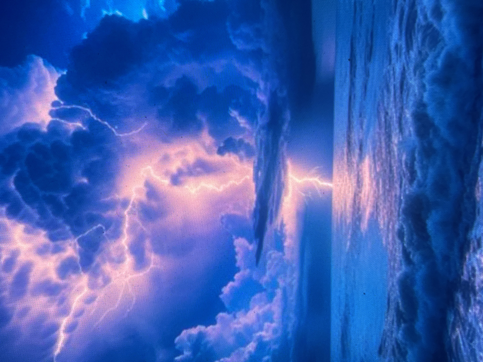 The image depicts a violent lightning storm over a turbulent body of water, likely an ocean or sea. The sky is filled with heavy, dark blue and purple storm clouds, some illuminated from within by the electric discharge. A prominent, bright pink-magenta lightning bolt strikes the surface of the water in the central midground, creating a vivid reflection. Other fainter lightning flashes are visible across the upper left and central sky. The water in the foreground is a deep, agitated blue, with distinct whitecaps and visible wave formations, also reflecting the pinkish hue of the lightning. No people, boats, or other discernible man-made objects are present in the scene. The overall environment is one of raw natural power and dramatic weather.