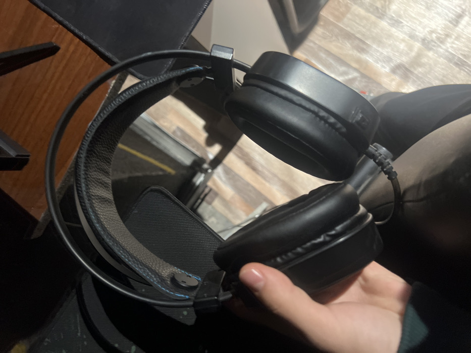 The image depicts a pair of black over-ear headphones being held by a human hand, observed in what appears to be an indoor setting in Tuyabuguz, Uzbekistan.
**Subjects/Objects:**
* **Headphones:** A pair of black over-ear headphones is the primary subject. The headband features a padded, textured strap with visible blue stitching along its edges. The earcups are black and appear to be made of a synthetic leather-like material. A black braided audio cable is visible extending from the left earcup.
* **Hand:** A light-skinned human hand, with clean, unpolished fingernails, holds the left earcup of the headphones. A dark green fabric, possibly a sleeve, is visible at the wrist.
**Actions/Interactions:**
* The hand is actively grasping the left earcup of the headphones, holding them upright.
**Environment:**
* **Background:** The floor visible in the background consists of wooden planks in varying shades of brown and beige. Dark, angular structures, consistent with furniture legs or undersides of a desk/table, are visible above the floor.
* **Lighting:** The scene is moderately lit, with shadows suggesting a directional light source, possibly overhead. The overall impression is an interior space.