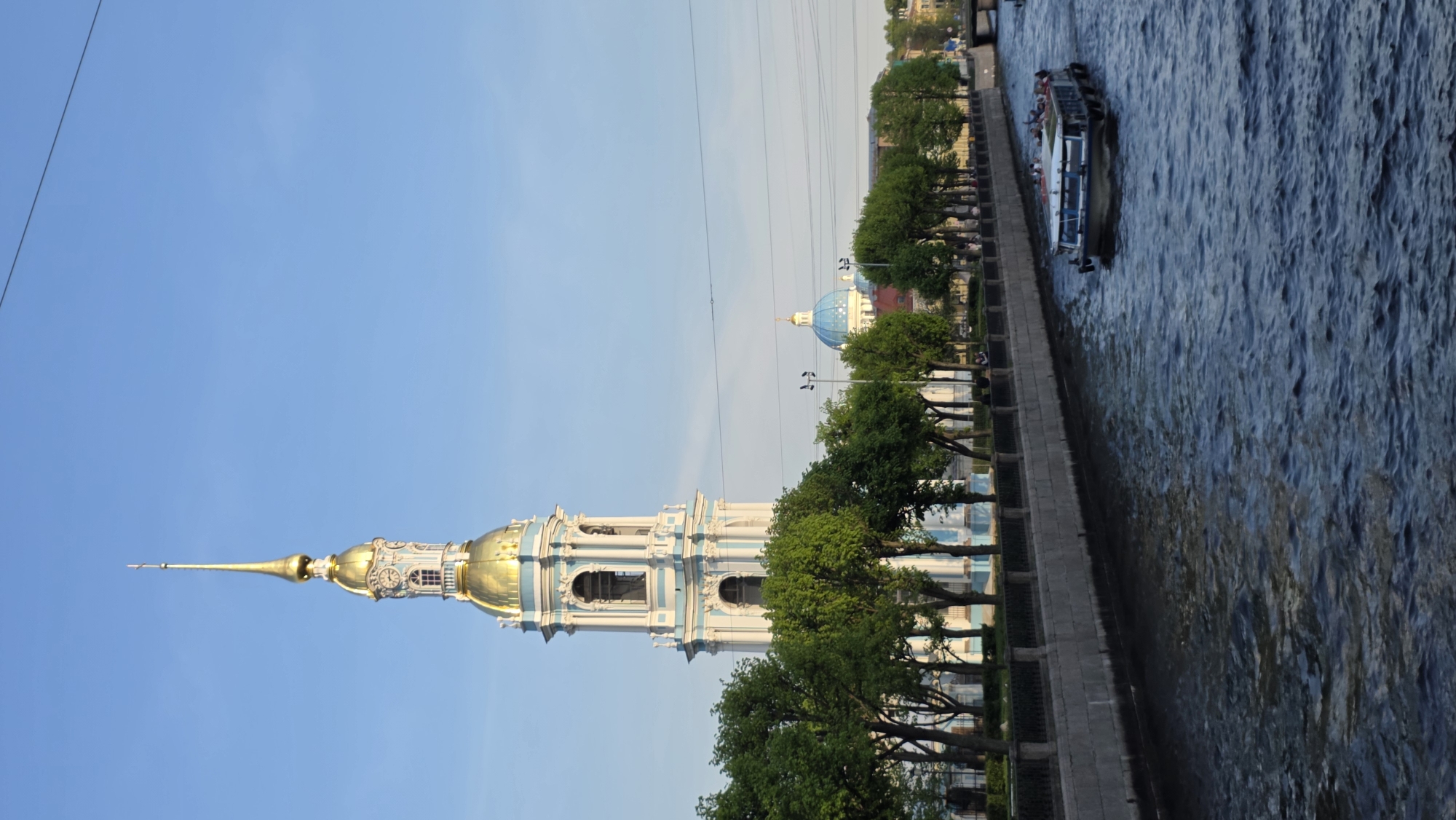 The image shows a view of Saint Petersburg, Russia, featuring the Naval Cathedral of Saint Nicholas with its gilded spire against a clear blue sky. A building with a blue dome can be seen farther down the river. Trees line the river bank. A boat with people on board is on the river. A stone embankment runs along the edge of the river. Power lines traverse the sky.