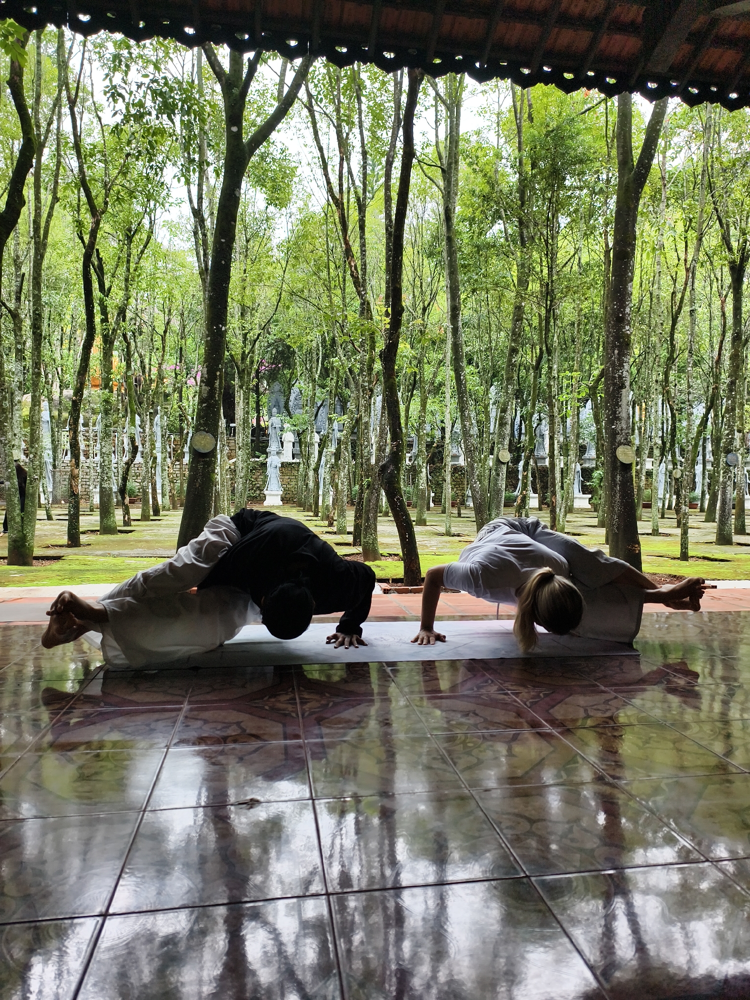 Two individuals are practicing yoga on a mat within a tiled, covered space, possibly a porch or pavilion. The individual on the left wears dark clothing, while the person on the right is dressed in lighter attire. Both are in a handstand position with their legs bent. Beyond the covered space, a dense grove of trees is visible, appearing to be part of a landscaped garden or park. Interspersed among the trees are light-colored, statue-like objects. The location is Unknown, Unknown.