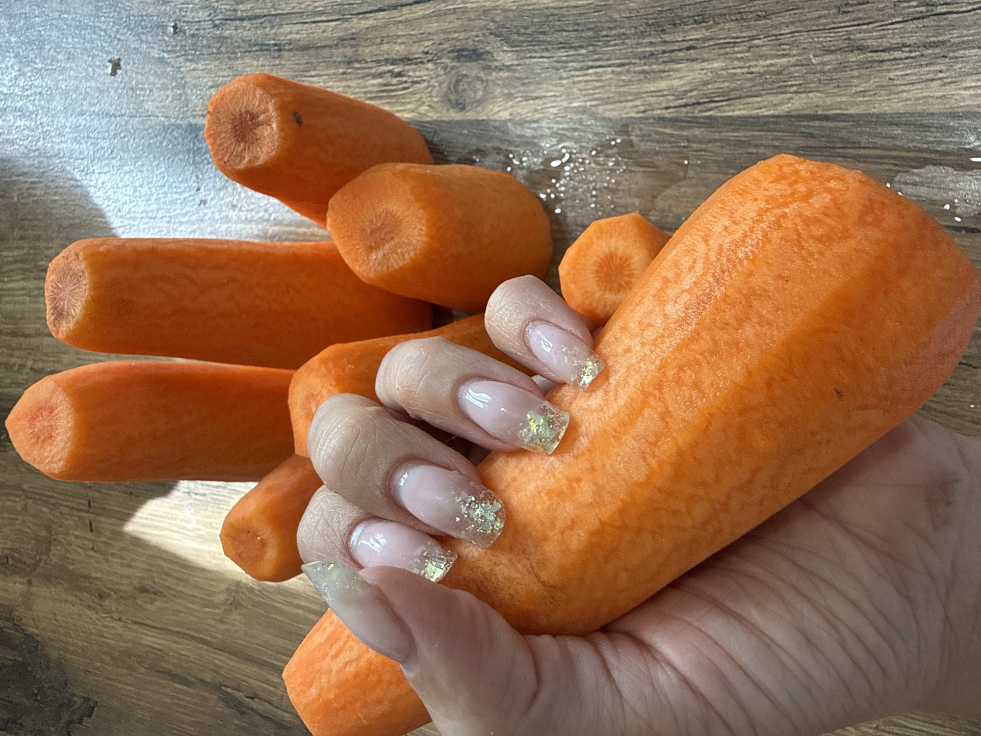 The image depicts a hand holding several peeled carrots on a wooden surface. The carrots are various sizes and lengths. The hand has long, manicured nails with a light pink base and glitter accents. There are water droplets on the wooden surface. There is no information to indicate the location is Заволжье, Russia.