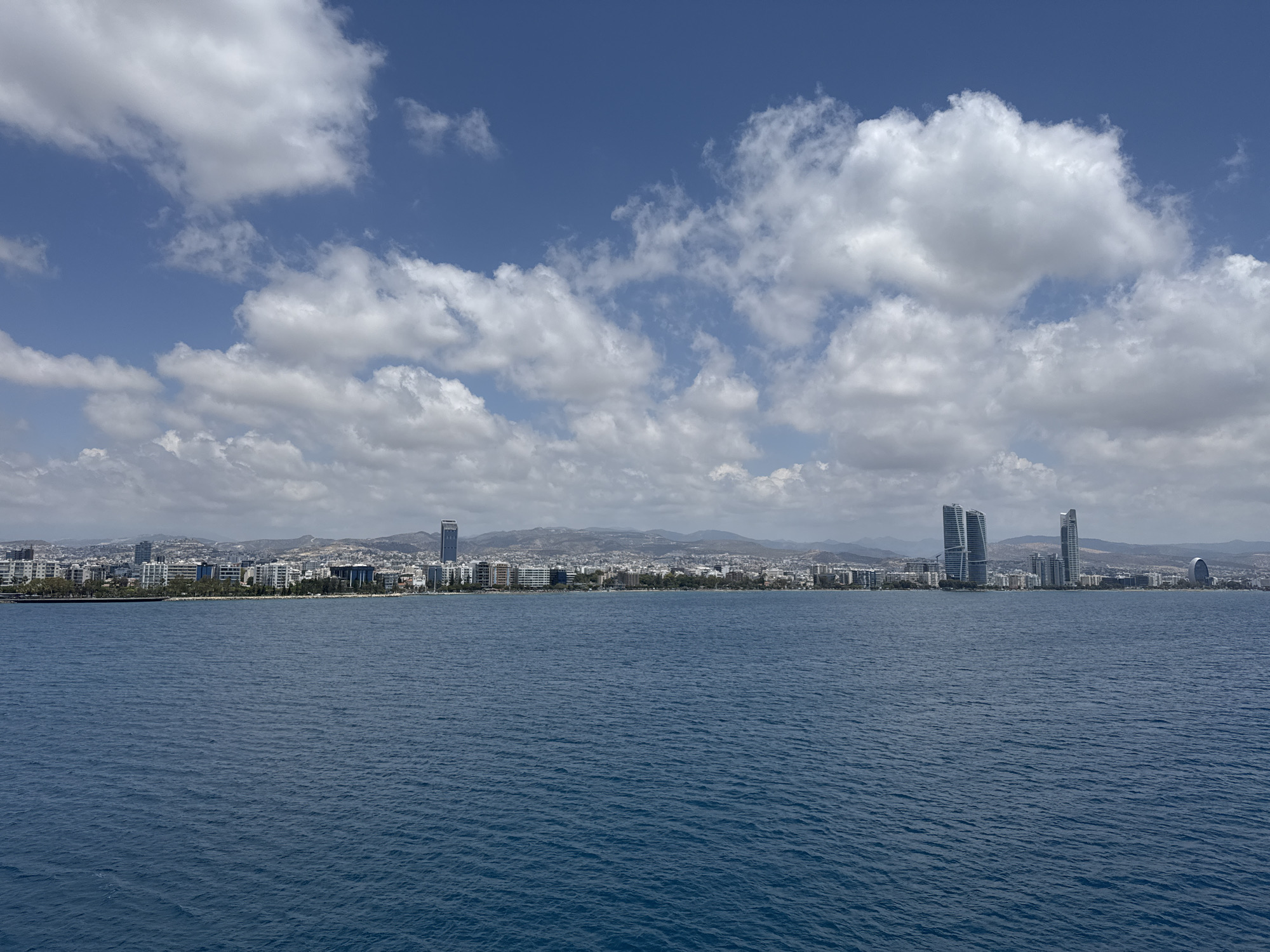 A wide, stable, horizontal shot captures a view of the coastline of Limassol, Cyprus, from the sea. The foreground is dominated by deep blue water with a slight ripple texture. The middle ground presents a long stretch of the city's skyline, featuring numerous buildings of varying heights and architectural styles, interspersed with trees. Prominent among the structures are several modern, tall skyscrapers, including two distinct, sleek towers that appear to be part of a business or residential complex. Further back, rolling hills form a backdrop to the urban sprawl. The sky above is a vibrant blue, populated by scattered white cumulus clouds. The overall impression is one of a sunny day, with the city extending along the coast under a partly cloudy sky.