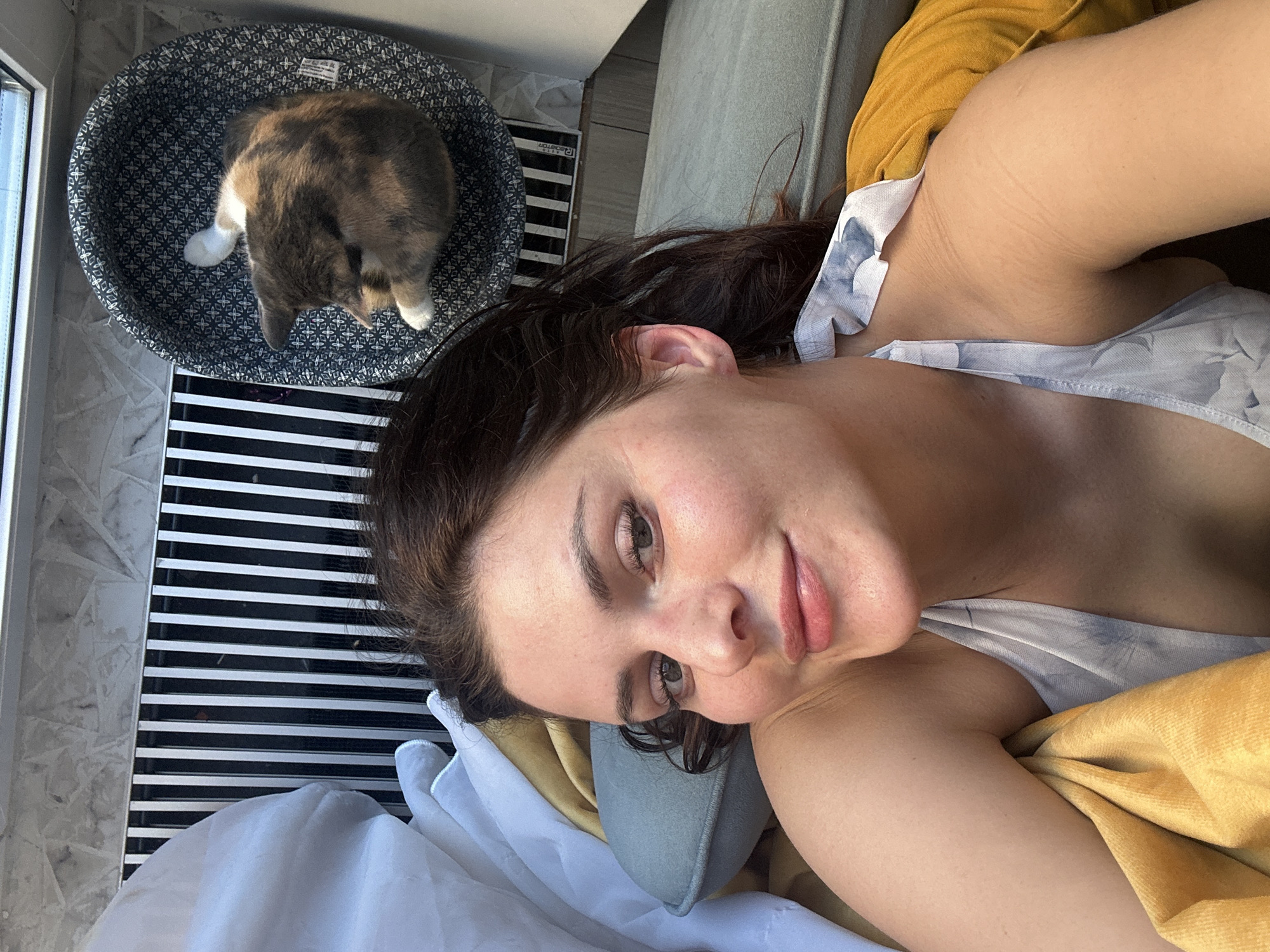 This media file is a selfie taken by a person lying down. The person is a young woman with dark hair and light skin. She is wearing a white top with a floral pattern. Her face is turned towards the camera, and she is smiling slightly. Her eyes are open and looking at the camera. The background is blurred, but it appears to be an indoor setting. There is a cat in a cat bed in the upper left corner of the image. The cat is a calico with patches of brown, white, and black. It is curled up in the bed, facing away from the camera. The lighting suggests it is daytime, possibly late afternoon or early evening, due to the warm tones. The overall mood of the image is relaxed and intimate. There is no visible text in the image.