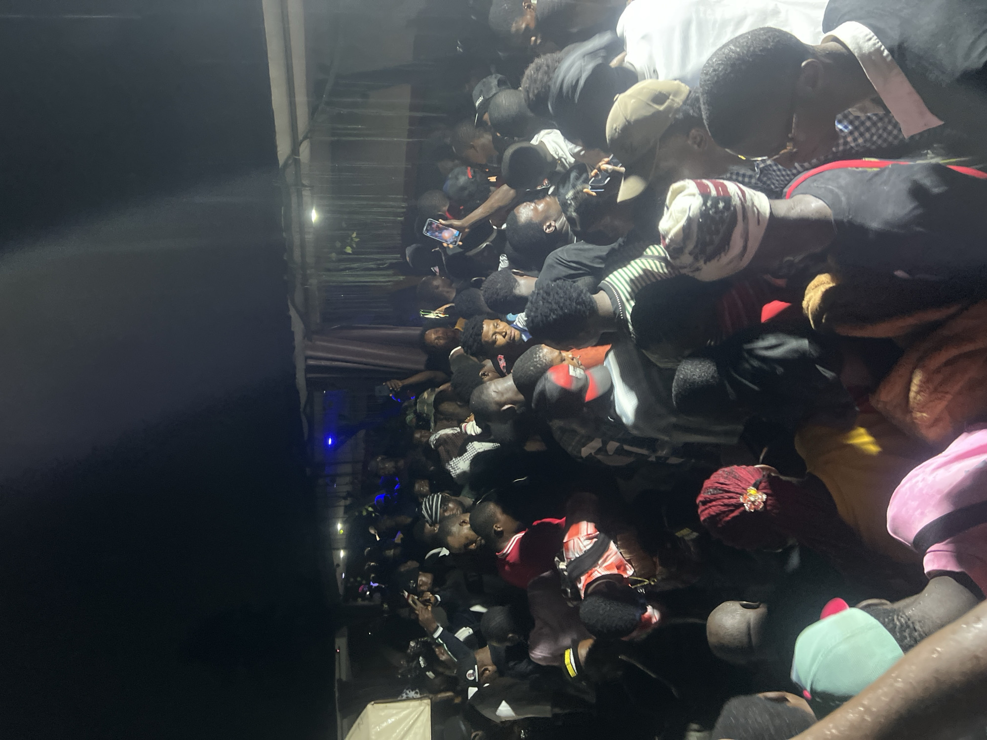 This is a nighttime, medium-shot photograph of a large gathering of people in Nkpor, Nigeria. The scene appears to be outdoors, possibly under a makeshift shelter, as evidenced by the corrugated metal roof structure visible at the top of the image. The lighting is dim, with a few bright artificial lights illuminating the crowd, creating strong shadows.
The crowd is densely packed, with individuals standing close together. Many people are facing forward or slightly to the side, suggesting they are observing something or someone in front of them. Some individuals are holding up their mobile phones, possibly to take pictures or videos. The attire of the people is varied, with many wearing casual clothing like t-shirts, caps, and headwraps. There are people of different ages and genders in the crowd.
The overall impression is that of a lively event, such as a concert, rally, or festival. The atmosphere seems energetic and engaged, with a sense of shared experience among the attendees. The presence of mobile phones indicates that people are documenting the event and possibly sharing it in real-time. The environment is somewhat rustic, consistent with an outdoor gathering in a less developed area. The dark sky and artificial lighting contribute to the nighttime ambiance.