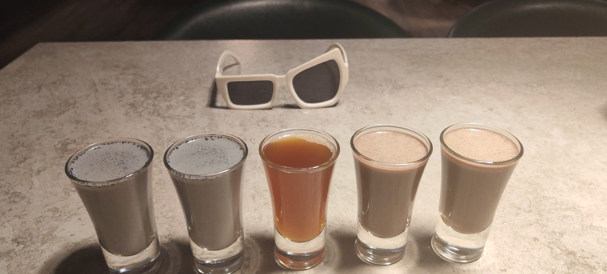 Five small glasses of drinks are arranged in a line on a speckled countertop. The leftmost two glasses contain a light gray liquid with dark specks on the surface. The third glass from the left holds a clear amber-colored liquid. The fourth and fifth glasses from the left contain a light brown, opaque liquid. Behind the glasses, a pair of white, angular sunglasses with dark lenses are resting on the countertop. The background is blurred, showing dark tones, suggesting an indoor setting. The image does not provide any information about the location in the city of Kaluga, Russia, or any people or actions.