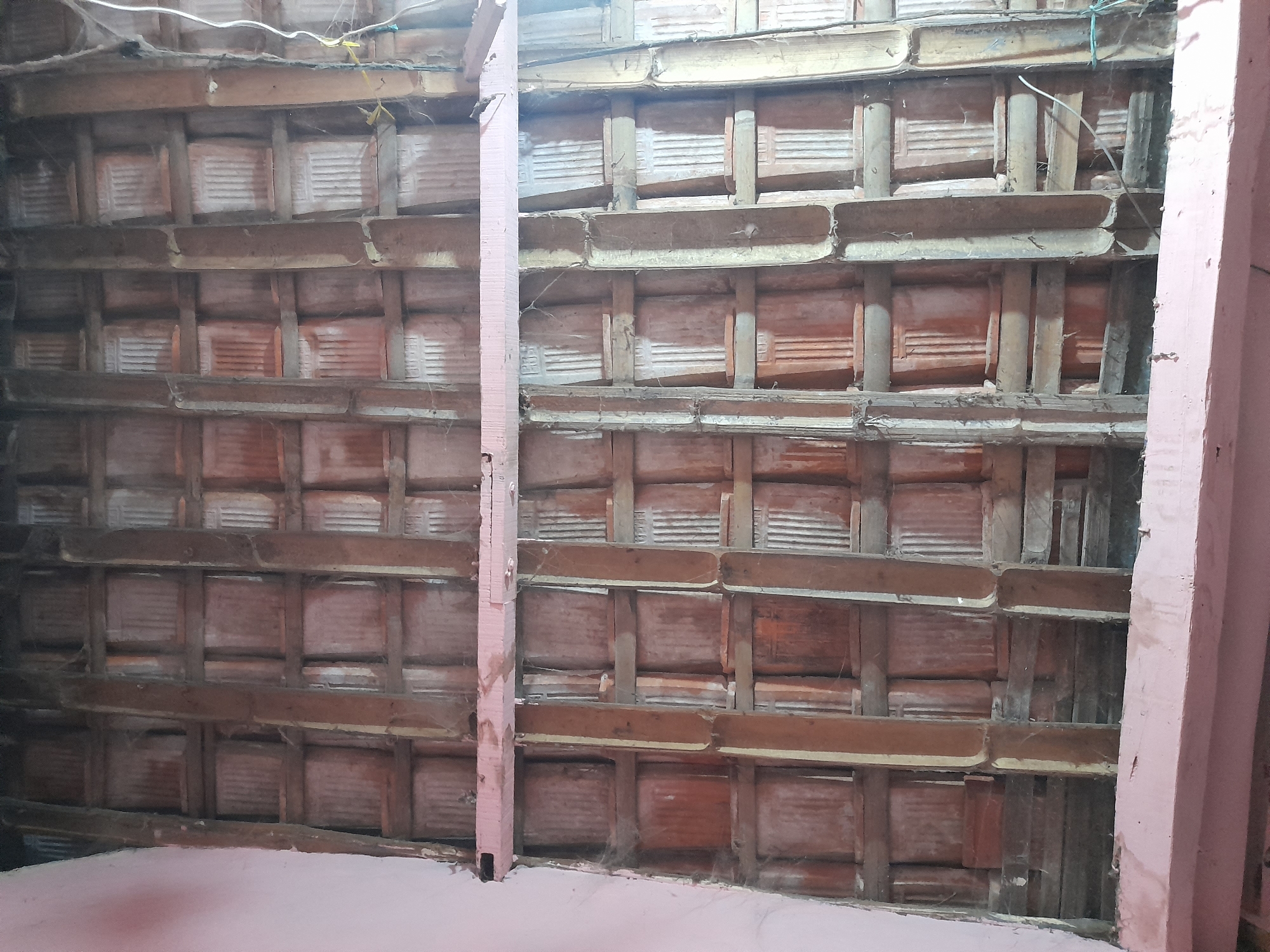 The image shows the underside of a damaged wooden roof structure with terracotta tiles. The roof appears to be in a state of disrepair, with some tiles missing and the wooden beams showing signs of age and wear. The scene is likely an interior space, possibly a room or attic, given the presence of what looks like a floor or lower ceiling in the foreground, painted a light pink color. Two vertical pink-painted wooden posts are visible on the right and left edges of the frame, supporting the roof structure or part of the building. Cobwebs are visible in several areas, indicating the space has not been recently cleaned or maintained. There are also electrical wires running across the top of the roof structure. The overall impression is one of an old, possibly abandoned or under renovation, building. The lighting is dim, suggesting it is either daytime with limited natural light or artificial lighting is being used. No people are visible in the image. No text is legible. The location context of Pulokulon, Indonesia, suggests a tropical climate where terracotta tiles are common roofing materials.