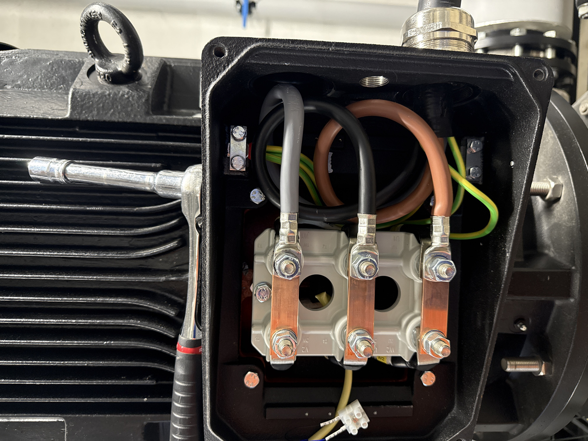 The image shows a close-up view of the internal wiring of an industrial electric motor. The motor casing is black and ribbed, with a lifting eye visible on the right side. A ratchet wrench is resting on top of the casing. Inside the motor's junction box, three sets of copper busbars are connected to electrical wires. Various cables, including brown, black, and grey, are visible, along with green and yellow striped grounding wires. The wiring appears to be neatly organized and secured. A white connector block with blue and yellow wires is also present. The scene is clean and well-lit, suggesting an industrial or workshop environment. No specific location in Antwerp, Belgium, can be determined from the image.