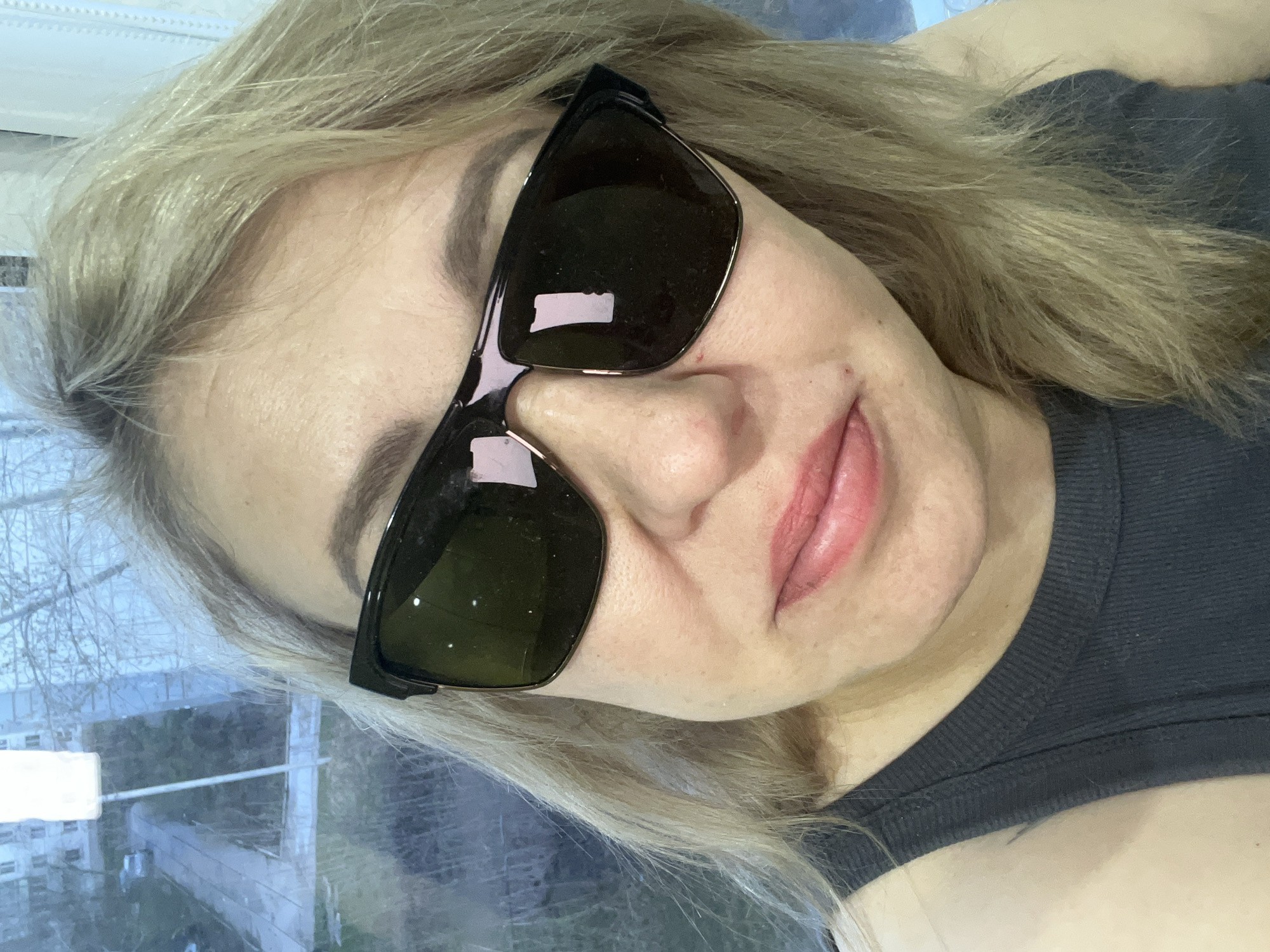 The image is a selfie of a woman with blonde hair, wearing sunglasses. She is wearing a gray tank top. A window is visible in the background, showing a view of what appears to be a residential area. Based on the prompt information, this is in Chaikovsky, Russia. Buildings and trees are visible through the window.
