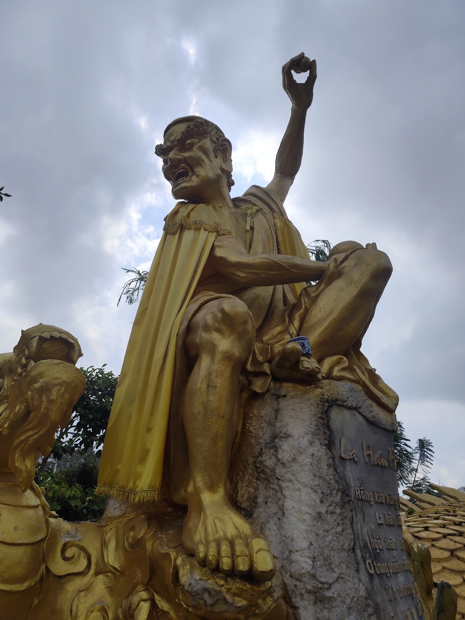 The image shows a golden statue of a seated figure with a raised right arm holding a small round object. The figure is draped in a golden cape. The statue is seated on a stone structure with Vietnamese text inscribed on it: "La Hán". Below the figure is a golden dragon-like structure. The sky is overcast with grey clouds. The location is Cam Ly - Đà Lạt, Vietnam.