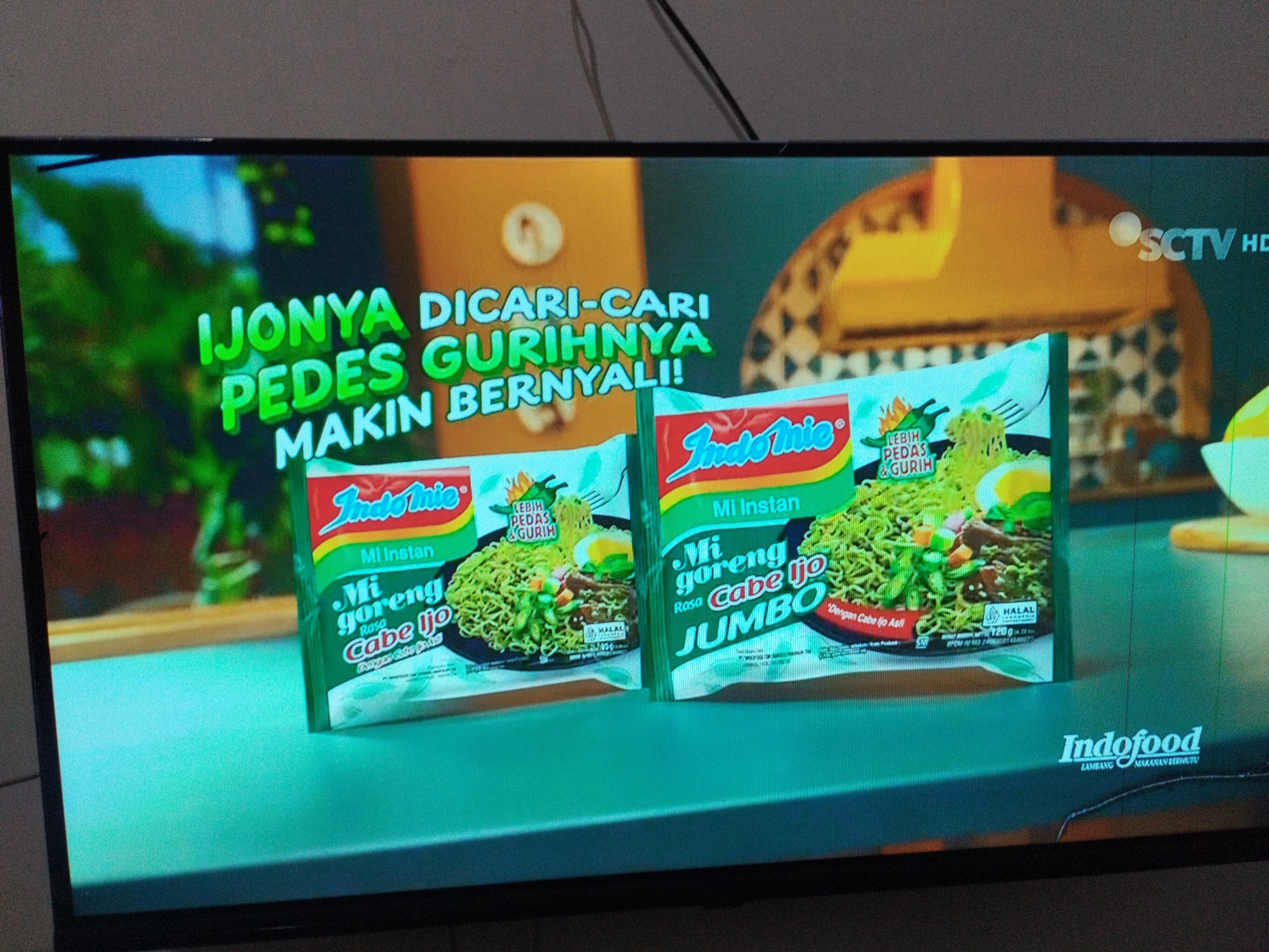The image shows a television screen displaying an advertisement for Indomie instant noodles. The advertisement features two packs of "Mi Goreng Cabe Ijo" flavor, one of which is a "Jumbo" size. Text on the screen reads "IJONYA DICARI-CARI PEDES GURIHNYA MAKIN BERNYALI!" which translates to "The craving is sought-after, its savory spiciness is getting bolder!". The Indofood logo is also visible. The background of the advertisement is abstract, with yellow and green elements. The image is a photograph of the television screen itself, and there are no visible people or specific city locations within the depicted advertisement or the photograph.