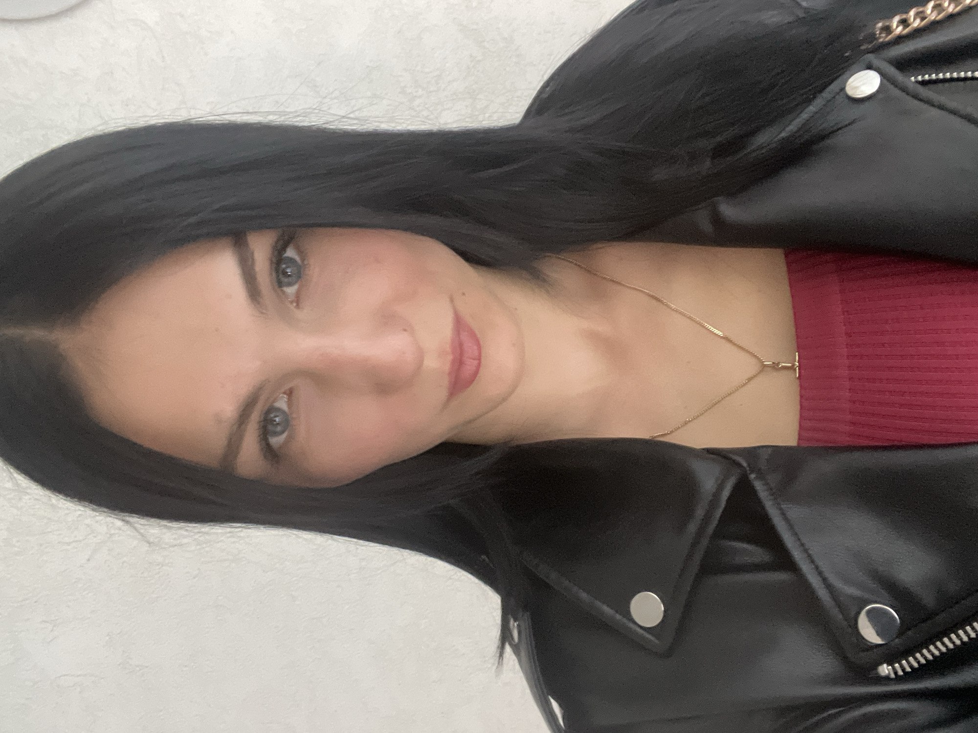 A close-up, medium shot shows a young woman with dark hair and blue eyes wearing a black leather jacket and a dark pink top. She is looking directly at the camera with a neutral expression. The background is a plain, light-colored textured wall.