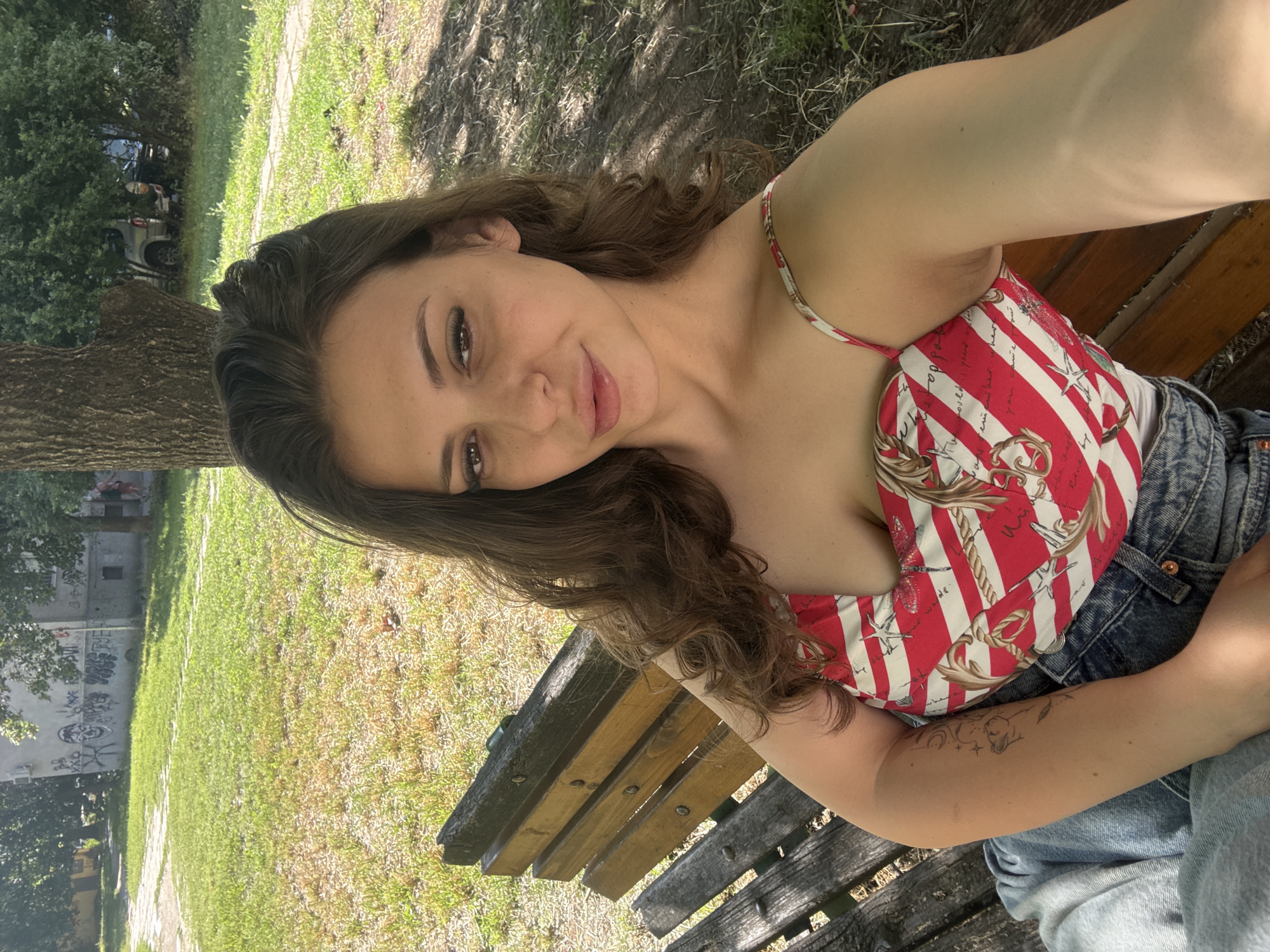 A young woman with long, wavy brown hair takes a selfie while sitting on a wooden park bench. She is wearing a red and white striped top with a nautical print and blue denim shorts. Her left arm is extended, holding the camera. She has a slight smile on her face and is looking directly at the camera. Visible tattoos adorn her left arm.
The setting appears to be a park with a grassy area and trees providing shade. The ground is covered in grass and dirt, with some fallen leaves. In the background, a paved path can be seen, as well as a building with graffiti on its exterior. The lighting suggests it is daytime, likely a sunny day, casting dappled shadows from the trees. The overall mood is relaxed and casual.