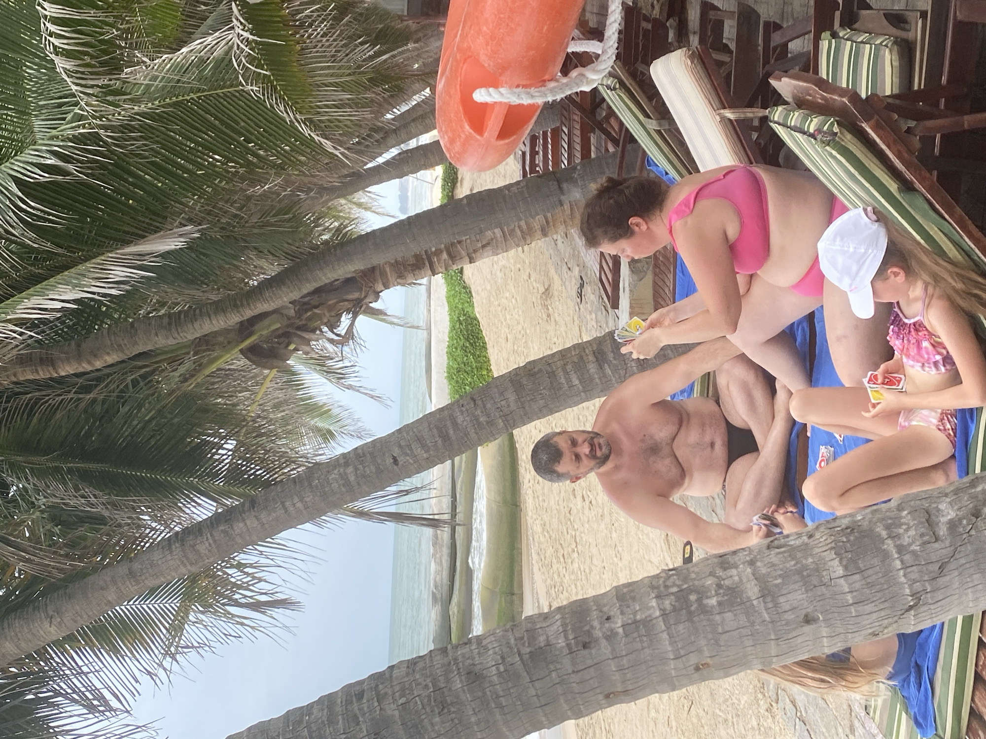 The image depicts a family playing cards on a beach in Mũi Né, Vietnam, amidst palm trees. A man, wearing black swim trunks, sits on a blue towel, facing the camera with a slight smile. Next to him, a woman in a pink bikini is also seated, holding a hand of cards and looking down. A young girl, wearing a pink and white patterned swimsuit and a white cap, is positioned to the right of the woman, also holding cards. Several chairs and a table with an orange flotation device are visible in the background, suggesting a beach resort setting. The scene appears to be outdoors on a sandy beach with the ocean visible in the distance. The light suggests it is daytime, and the weather seems pleasant, as people are dressed in swimwear. The overall mood seems relaxed and casual.