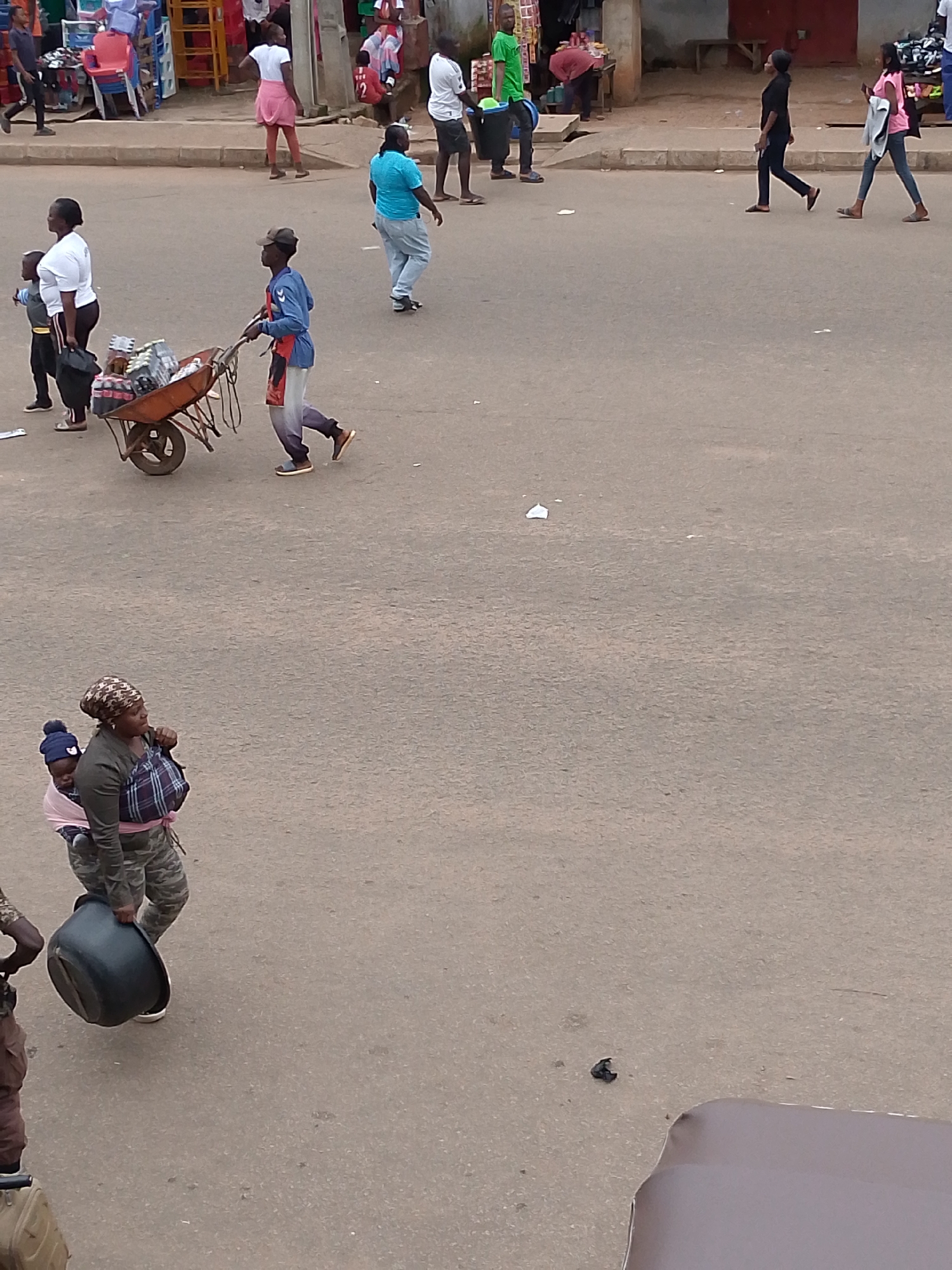 This is a high-angle, medium shot capturing a street scene in Bukuru, Nigeria, with a focus on pedestrian activity and market stalls. The pavement is a dull, uneven gray, with scattered debris and occasional lighter patches. On the left, a woman with a headscarf carries a baby strapped to her back, also holding a large, dark basin in her right hand. She wears camouflage pants and a plaid cloth around her waist.
Further up the street, a young man pushes a wheelbarrow filled with goods, some appearing to be bottled beverages. He is dressed in a blue long-sleeved shirt, a red apron, and light-colored pants. To his left, a child in a white shirt and dark shorts walks alongside a woman in a white shirt and black pants, who is also holding a black bag.
In the background, a vibrant collection of market stalls and seating arrangements is visible. Red and blue plastic chairs are stacked, and various goods are displayed on tables and shelves. Several people are seen walking and interacting around these stalls. A man in a green shirt carries a blue bucket, and others are dressed in casual attire. The overall atmosphere suggests a bustling local market environment. The buildings in the background are simple structures, some appearing to be made of concrete.