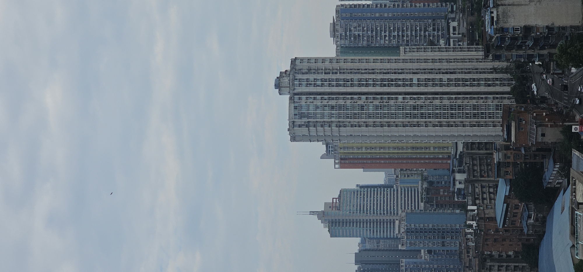 An aerial vertical outdoor shot captures a dense urban landscape in the Jiulongpo District, China, under a cloudy sky. The scene is dominated by numerous high-rise apartment buildings of varying architectural styles and colors, densely packed together. In the foreground, older, darker brick buildings with visible window units contrast with the sleek, modern designs of the newer towers. The middle ground features several prominent, tall buildings, including a light blue skyscraper with horizontal banding and a very tall, white, rectangular building with a grid of windows. Other buildings display splashes of color like red, yellow, and green. A winding road with several cars is visible at the bottom of the frame, suggesting ongoing city traffic. The sky is overcast, with visible clouds, and a single, dark, bird-like object is present in the upper left portion of the sky. The overall impression is one of a sprawling, heavily populated metropolitan area.