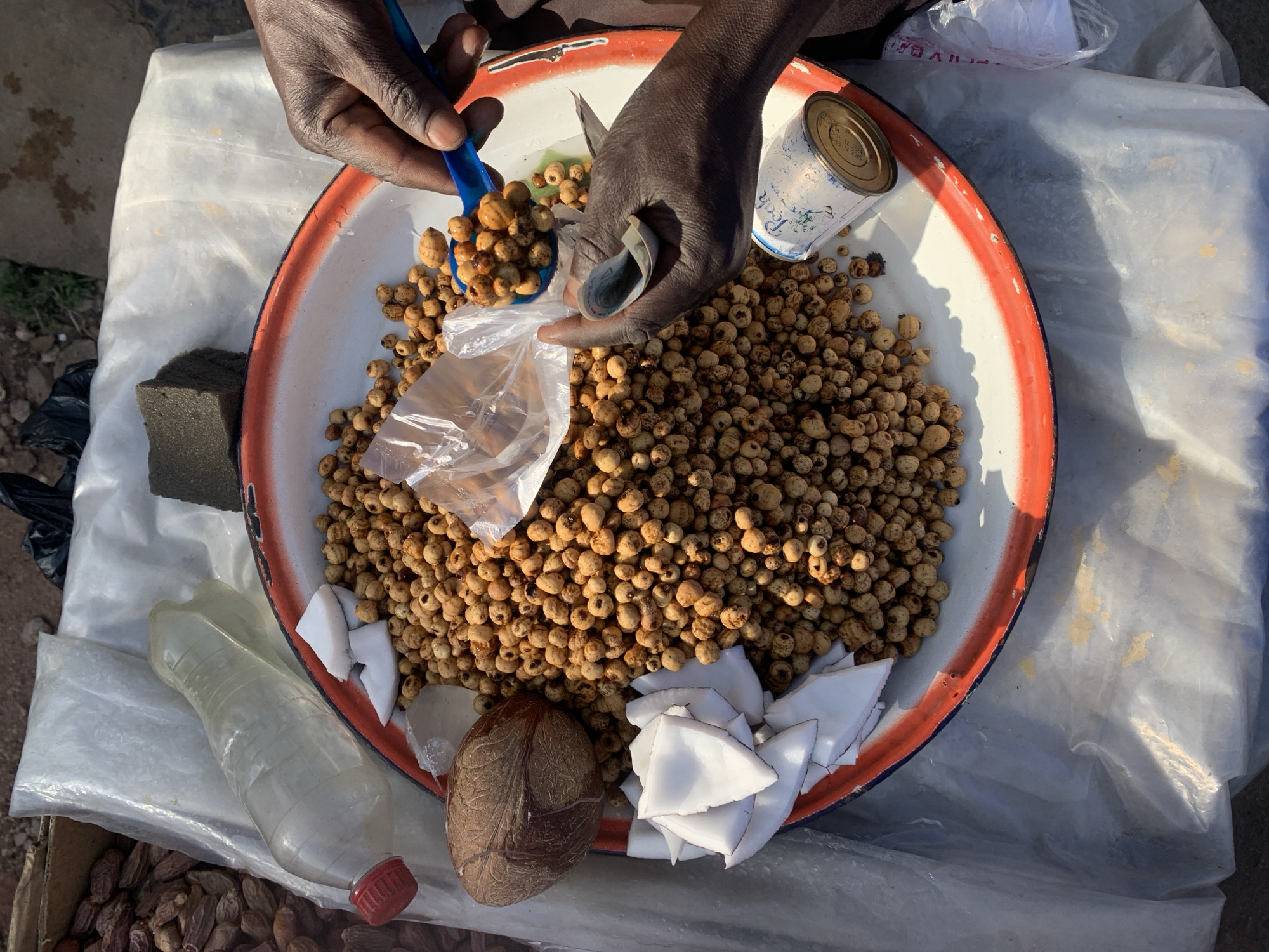 A vendor at New Stadium in Nigeria sells tiger nuts from a large metal pan. A person with dark skin uses a blue spoon to scoop tiger nuts into a clear plastic bag. The person holds a rolled-up bill. The pan also contains a halved coconut, chunks of white coconut meat, and a small can. A plastic bottle and a dark sponge are near the pan. The pan sits on a white plastic sheet.