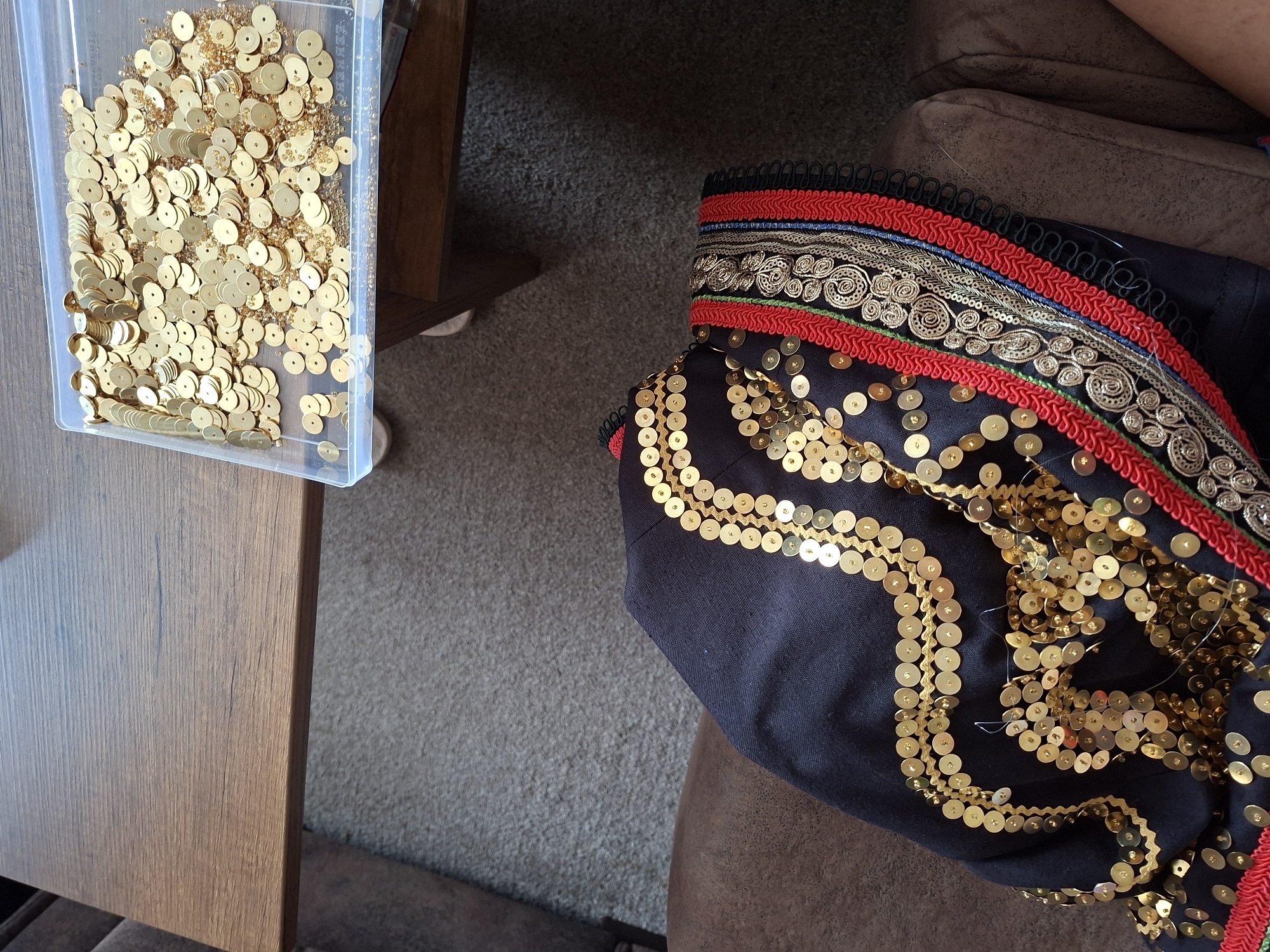 A close-up, high-angle shot captures a sewing project in progress. A dark navy garment, possibly a jacket or dress, is draped over the arm of a brown leather couch. The garment is heavily embellished with rows of gold sequins, some of which are sewn onto the fabric, while others are loose and scattered across the surface. A clear plastic container filled with loose gold sequins and small amber beads sits on the edge of a dark wood table, partially visible in the upper right of the frame. The table is positioned over a neutral-colored carpet. The scene appears to be indoors, likely in a home setting, with no identifying external environmental features.
