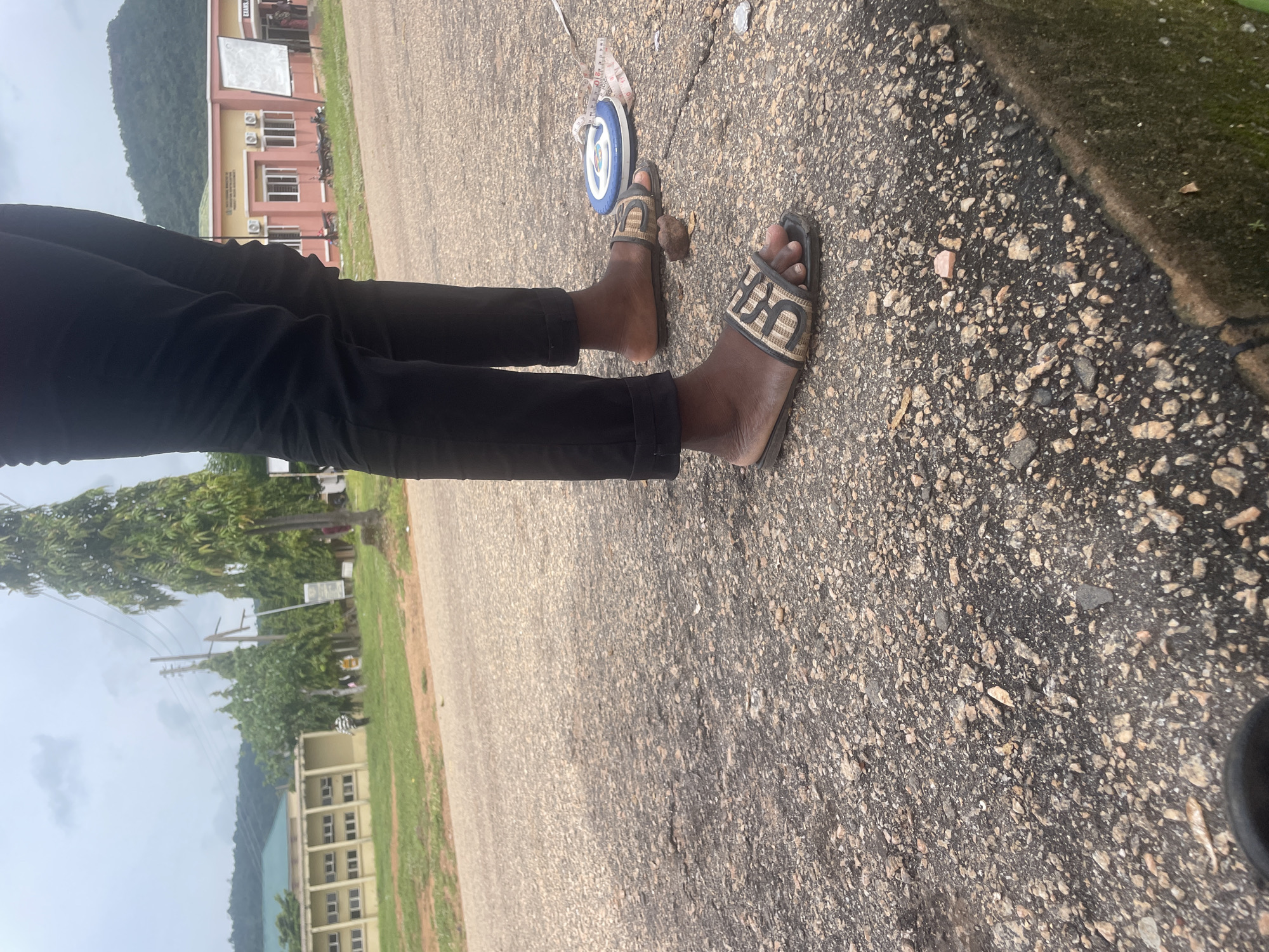 The media file shows a person's legs and feet wearing black pants and slide sandals, standing on an asphalt surface. In the background, there is a building with a light brown facade, multiple windows, and air conditioning units. Green trees and a hill are visible behind the building. A tape measure is lying on the ground near the person's feet, and there is a small, dark object, possibly a rock, also near the feet. The scene appears to be outdoors, with an overcast sky suggesting it could be a cloudy day. The location is identified as Lokoja, Nigeria. The overall impression is a casual outdoor setting, possibly on a campus or public area. There is no discernible text in the media file.