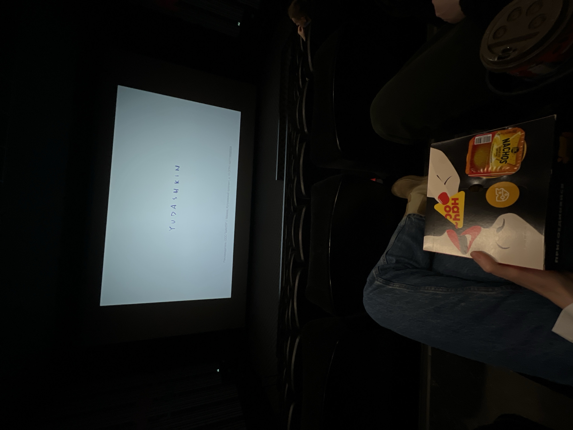 A person is sitting in a movie theater, holding a box of nachos and a drink. The movie screen shows the name "Yudashkin" with Russian text below it. The text suggests the movie is being shown in Moscow, specifically, at 10 Kutuzovsky prospect. The person is wearing jeans and a white sleeve of what appears to be a long-sleeved shirt. The location, based on the text on the movie screen, is a movie theater in Moscow.