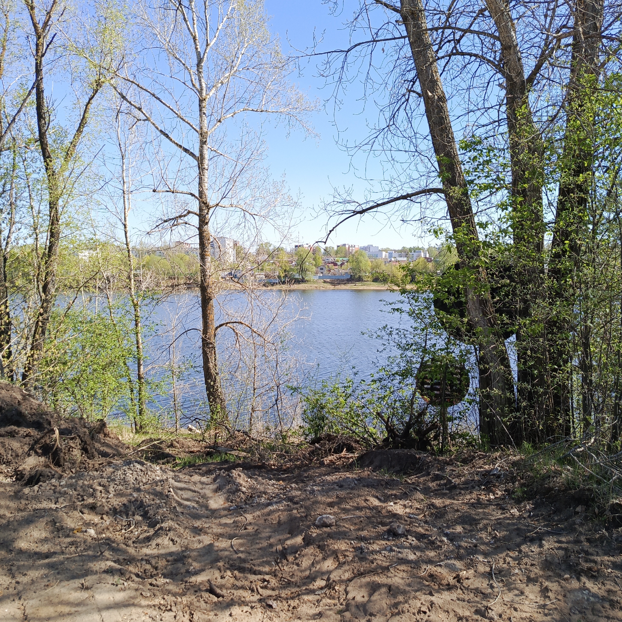 The image shows a dirt path leading down to the Votkinsk Reservoir in Chaikovsky, Russia. In the background, across the water, there are buildings of varying heights, a mix of residential and commercial structures. The sky is clear and blue. The trees on either side of the path are in the early stages of leafing out.