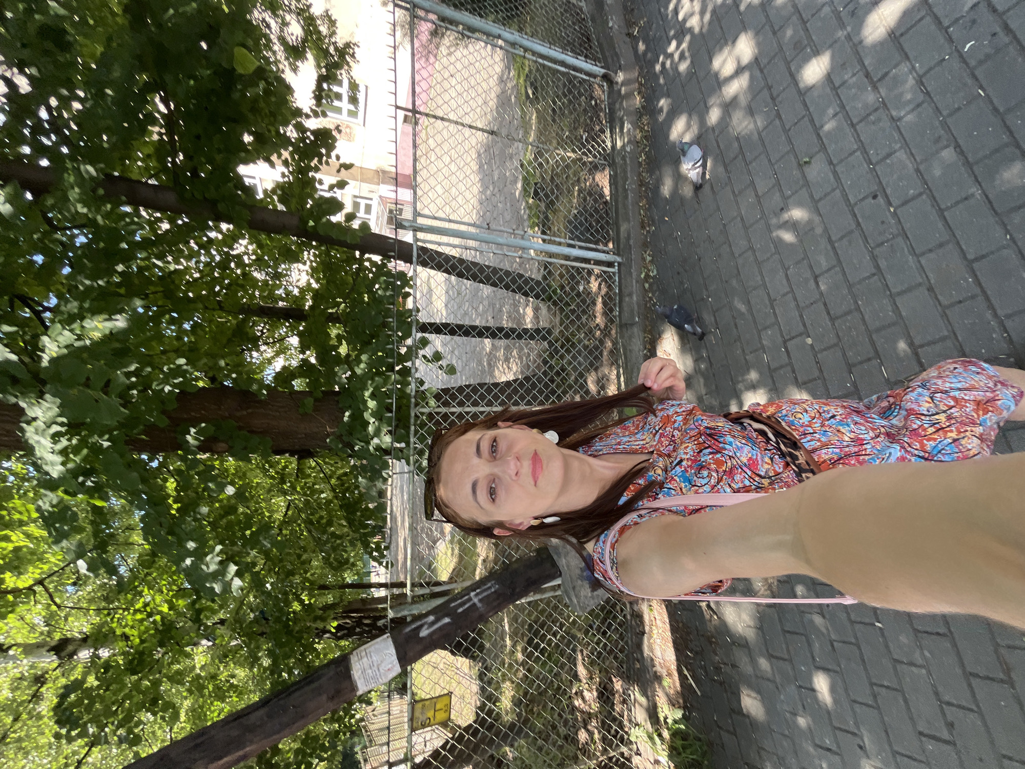 A woman is taking a selfie on a sidewalk in front of a chain-link fence and trees. She is wearing a colorful, patterned dress and has long brown hair. There are two pigeons on the sidewalk. In the background, there is a building and more trees. The sidewalk is paved with dark gray rectangular bricks. The sun is shining, creating dappled shadows on the ground. The setting appears to be an urban park or a street with trees.