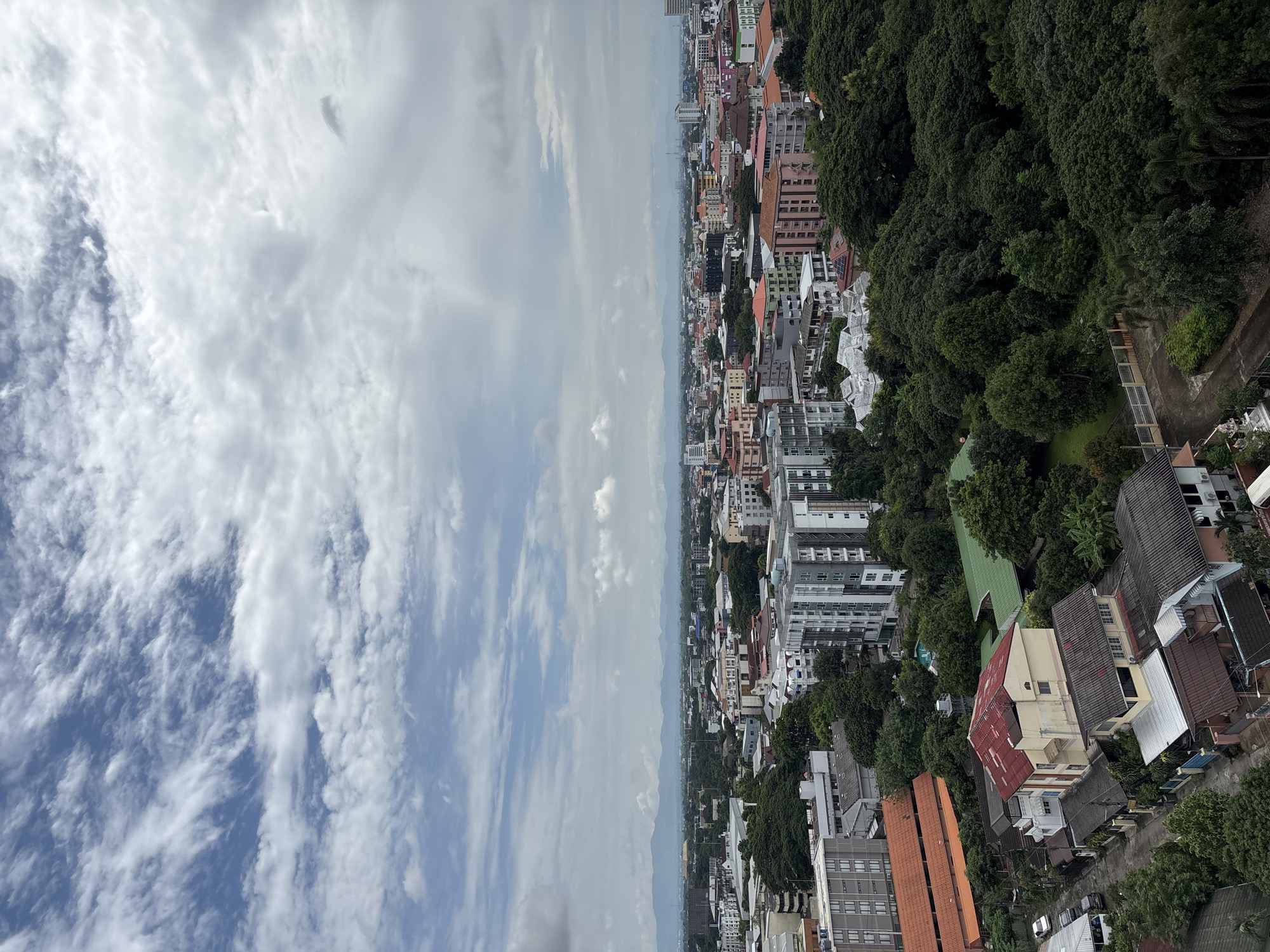 This is an aerial, wide-angle, daytime, eye-level view of a cityscape in Chiang Mai City Municipality, Thailand. The sky is overcast with scattered clouds, some of which are thin and wispy, while others are denser. In the foreground, there is a dense canopy of lush green trees interspersed with residential buildings and some taller apartment complexes. The buildings vary in architectural style and color, with some featuring orange, red, cream, and grey facades and roofs. Roads and parked vehicles are visible between the houses. In the middle ground, more apartment buildings and urban structures are present, with varying heights and colors. The background features a hazy, distant mountain range under the diffused sky. The overall impression is one of a densely populated urban area with significant green spaces. There are no people or animals visible in the frame.