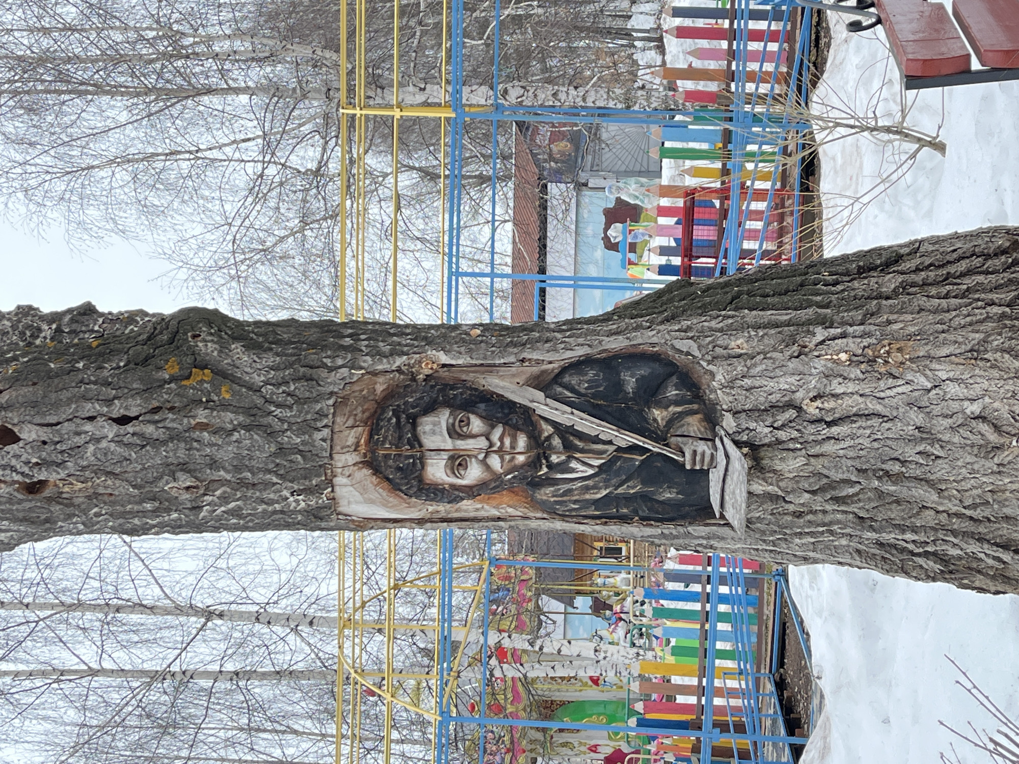 A portrait of a man is carved into the trunk of a tree. The man has dark hair and is holding a shovel. The tree is located in the city of Chaikovsky, Russia. In the background is a colorful playground and a bench. It appears to be winter with bare trees and snow on the ground.