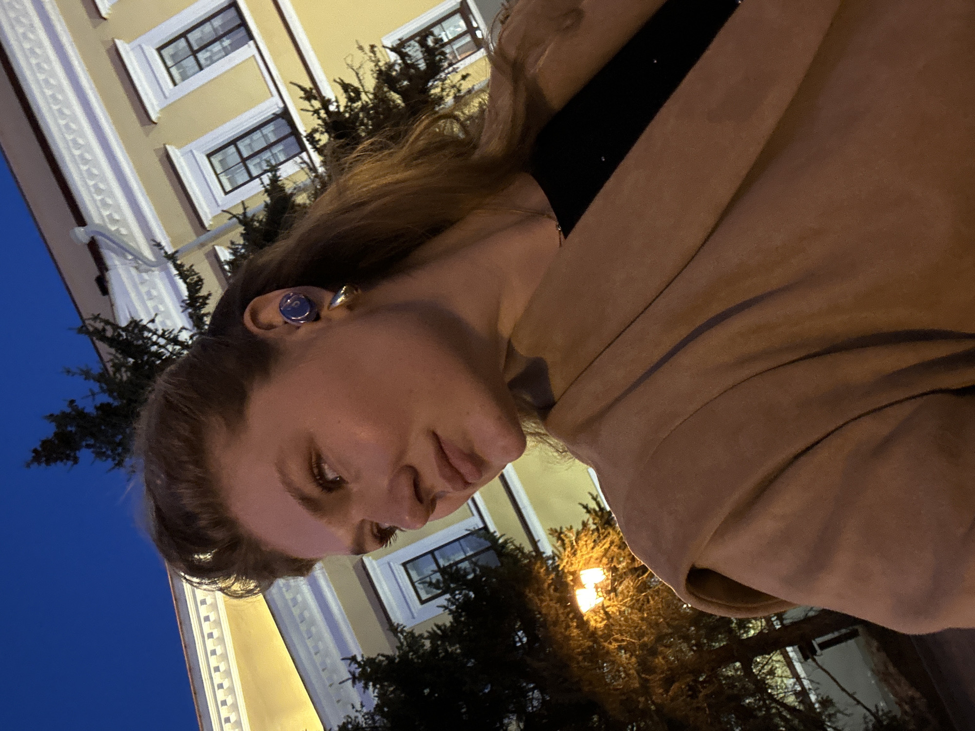 The image shows a woman wearing a tan jacket and a black shirt. She is standing outside in what appears to be Kazan, Russia, given the architectural style of the building behind her. The building is light yellow with white window frames and decorative white moldings. There are trees planted around the building and a street light illuminates them. The sky is dark blue, indicating it may be dusk or night. The woman is turned to the side, but looking down toward the camera with a slight smile. She is wearing an earring on her left ear.