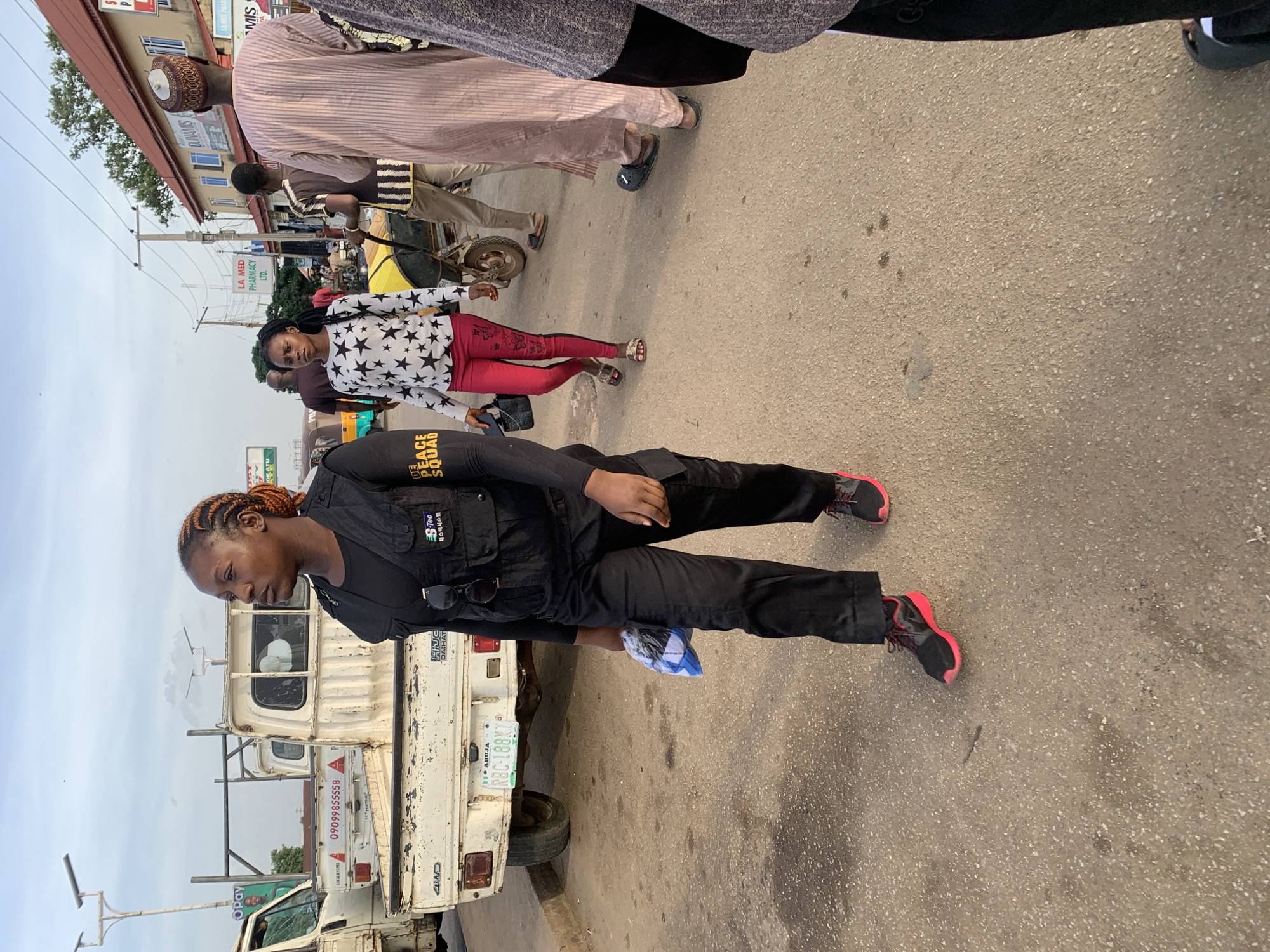 The scene is a street in what may be Gura Top, Nigeria. A woman in black clothing with a "PEACE SQUAD" patch stands near a white Daihatsu 4WD pickup truck with the license plate "RBC-188XT ABUJA." The truck has a phone number, 09099855558, displayed on it. She wears red and black sneakers. In the background, a woman in a star-patterned shirt and red pants walks on the street. Further back, a man leads a horse-drawn cart. Buildings with signs such as "LA MED PHARMACY LTD." and "DUNANIS" are visible in the background. A person wearing a traditional cap is seen in the upper portion of the picture.